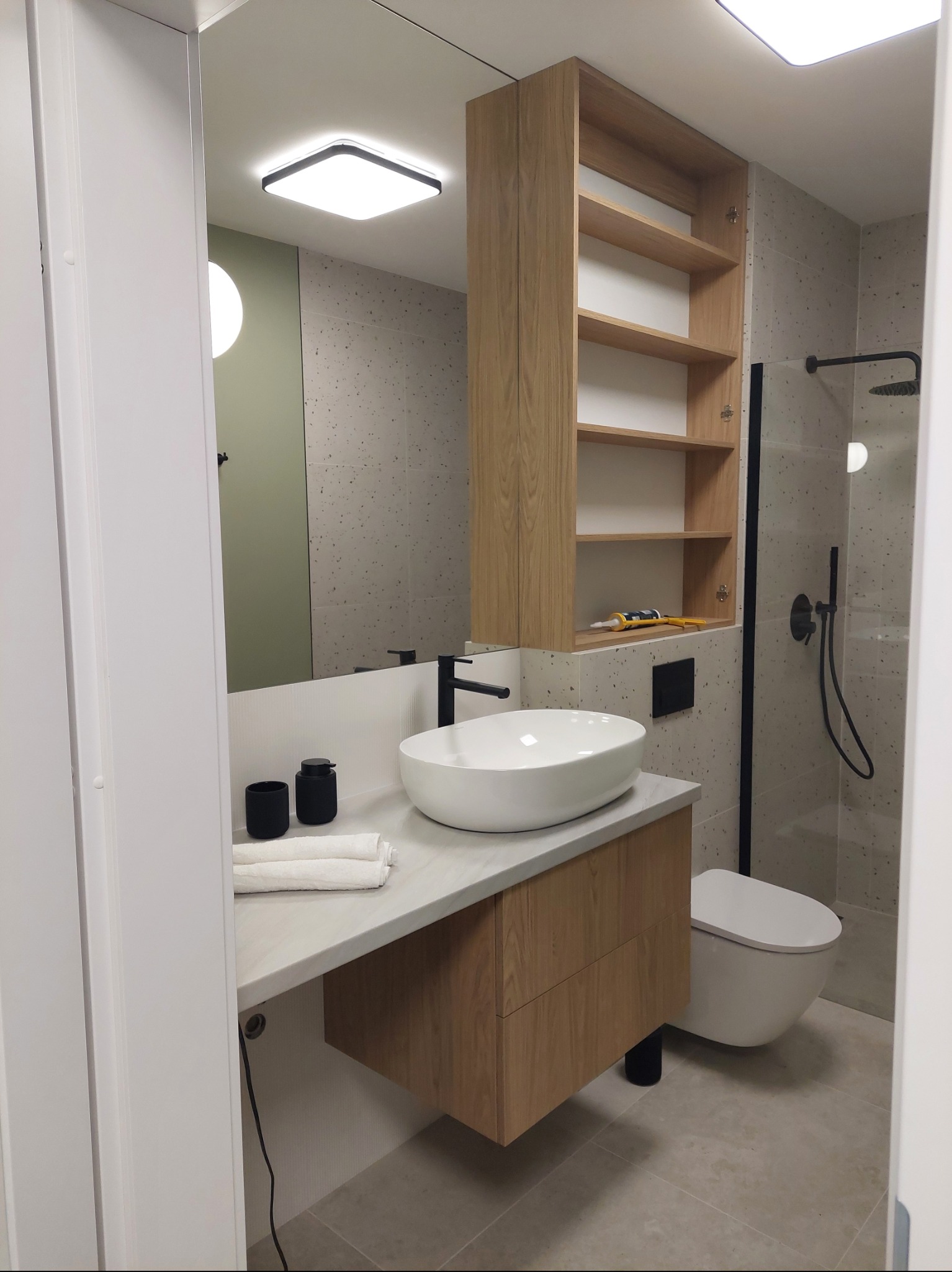 Nowoczesna łazienka z drewnianą szafką pod umywalką, lustrem i otwartymi półkami. Na półce tuba silikonu i szpachelka. Minimalistyczny design, płytki w odcieniach szarości.