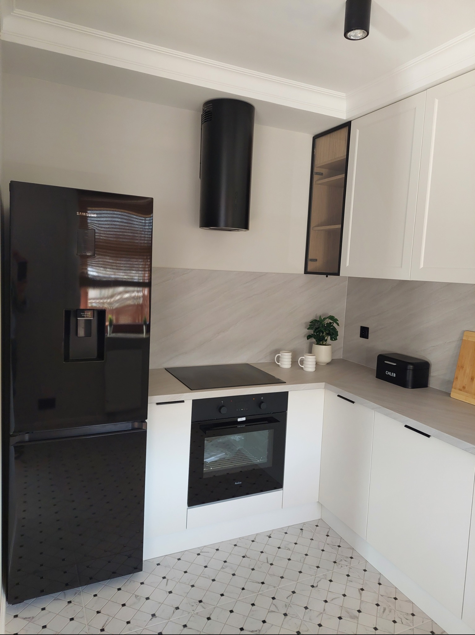 Elegancka kuchnia z czarną lodówką Samsung, zabudowaną piekarnikiem i białymi szafkami. Uwagę przykuwają marmurowe płytki podłogowe i minimalistyczny okap.
