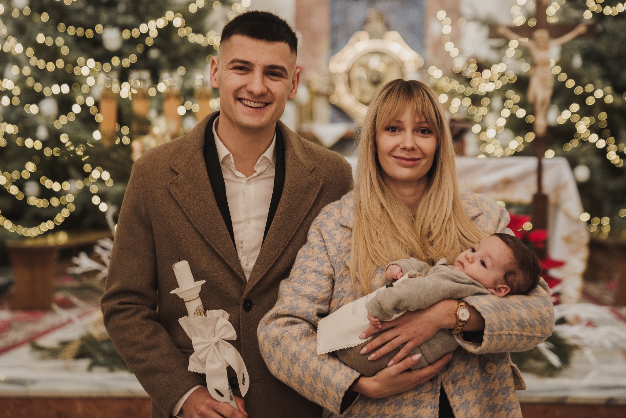 Uśmiechnięta rodzina z dzieckiem i świecą w kościele, dekoracje świąteczne w tle. Pamiątka z uroczystości religijnej.