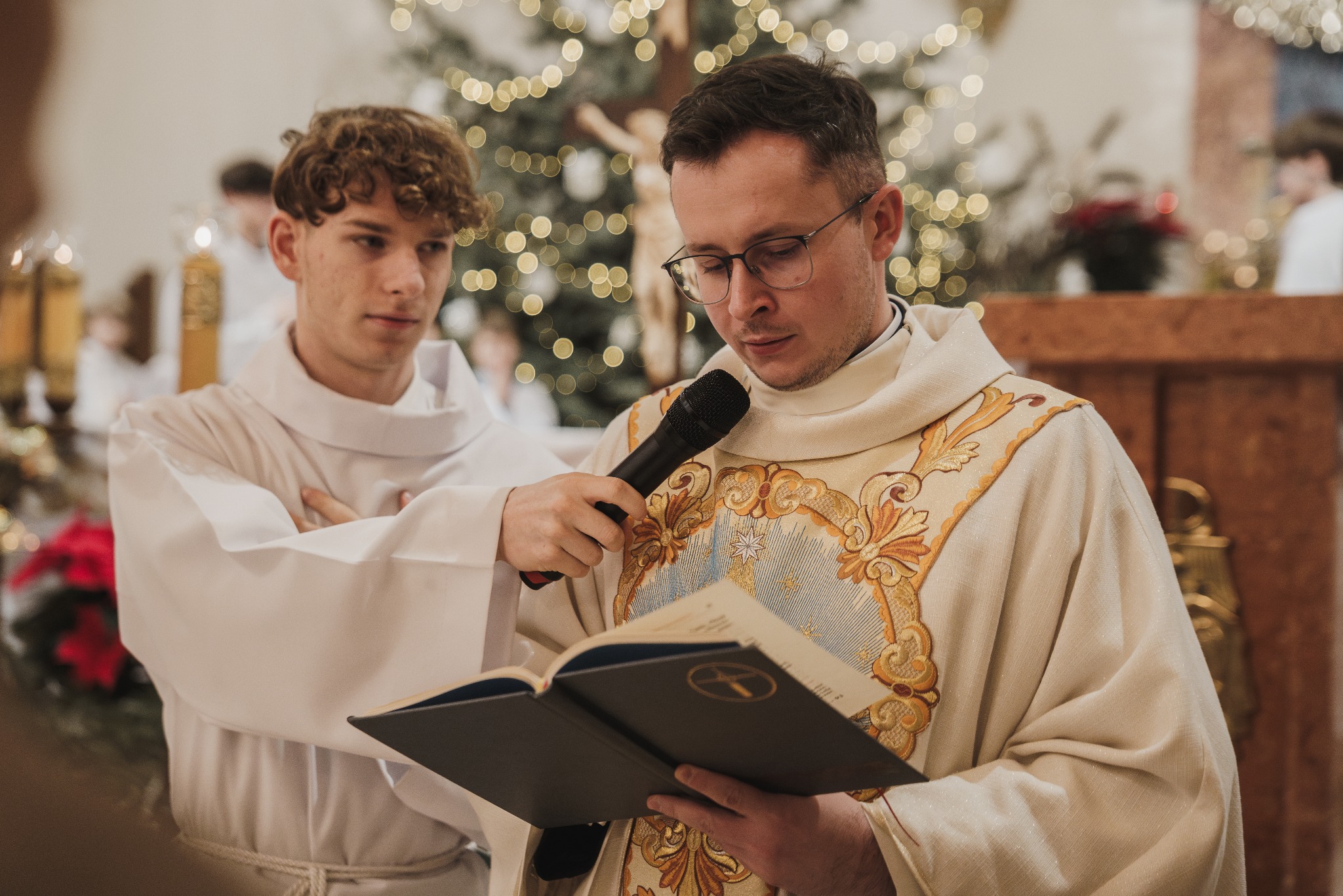 Ksiądz w bogato zdobionej szacie czyta z księgi do mikrofonu, obok ministrant. W tle oświetlona choinka i krucyfiks. Uroczysta atmosfera w kościele.