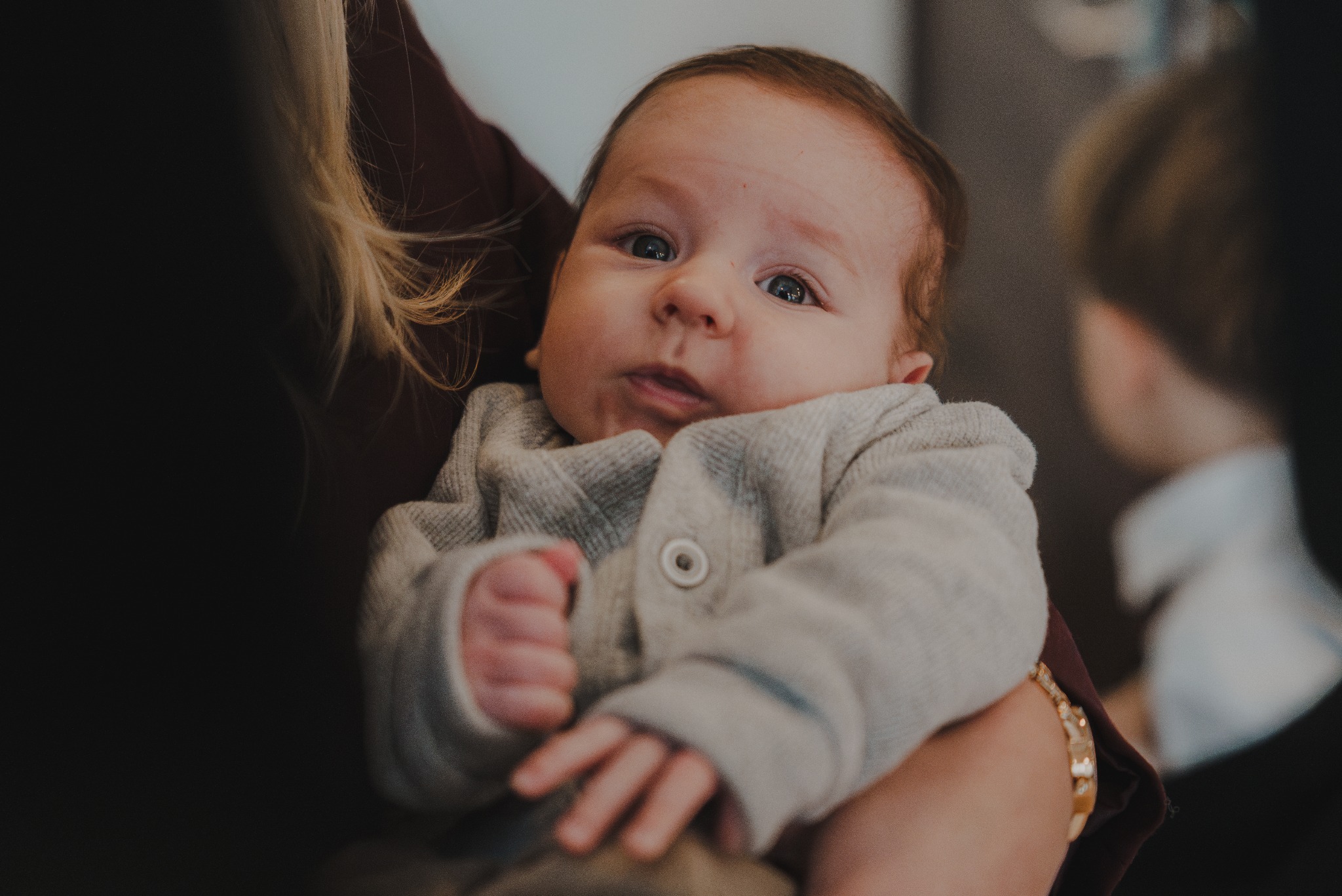 Portret niemowlęcia w szarym sweterku, trzymanego na rękach. Dziecko patrzy w obiektyw, ma jasną cerę i ciemne włosy. W tle widoczne fragmenty sylwetek innych osób.
