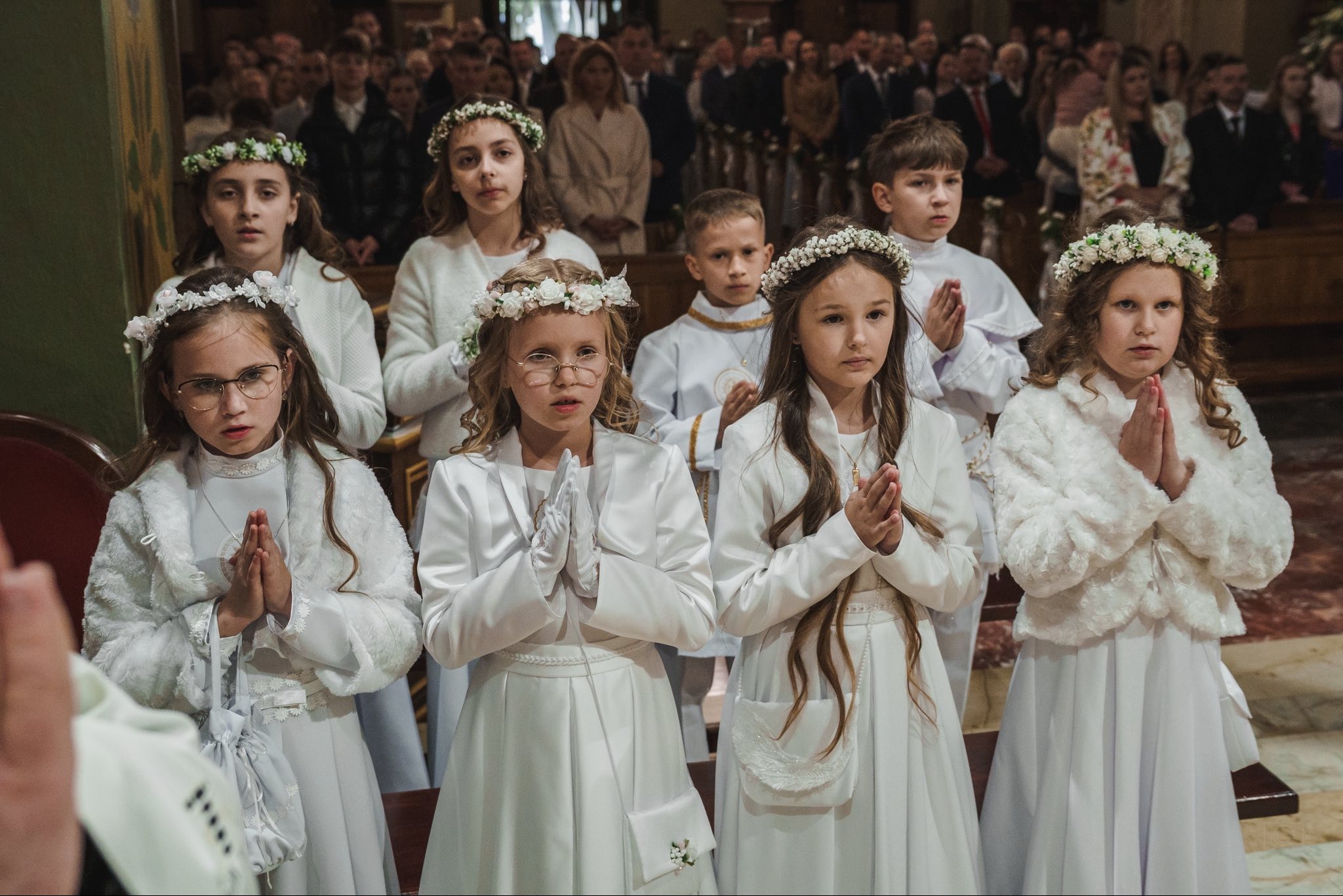 Uczestnicy Pierwszej Komunii Świętej w białych strojach i wiankach na głowach, stojący w kościele z złożonymi rękami do modlitwy, w tle widoczni inni wierni.