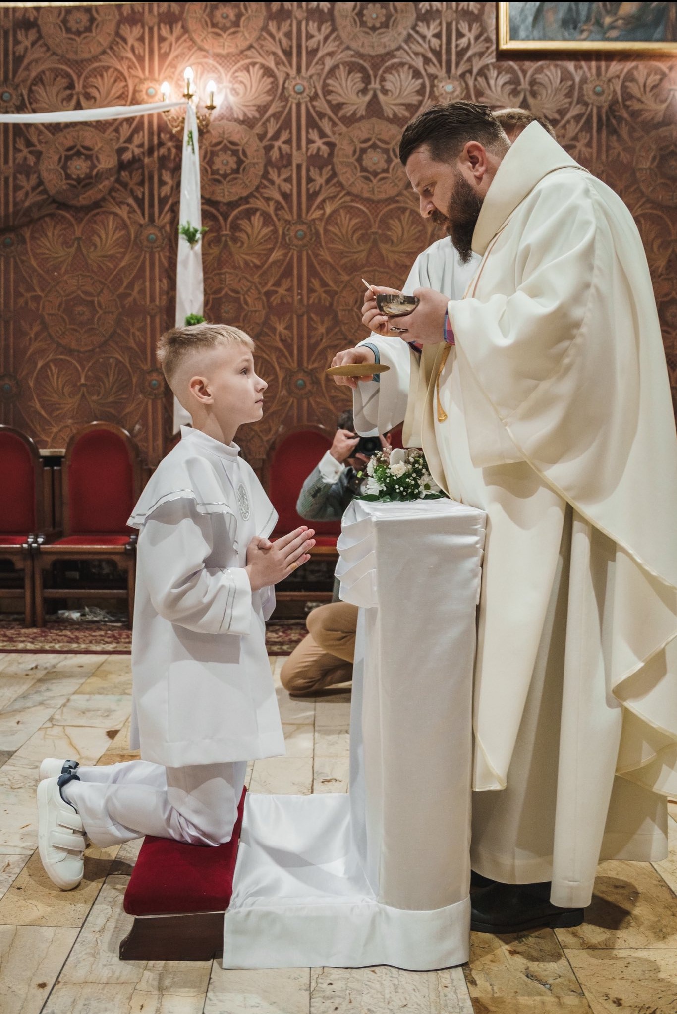 Chłopiec w albie klęczy podczas Pierwszej Komunii Świętej, przyjmując komunię od księdza w kościele. W tle dekoracyjne ściany i fotograf.