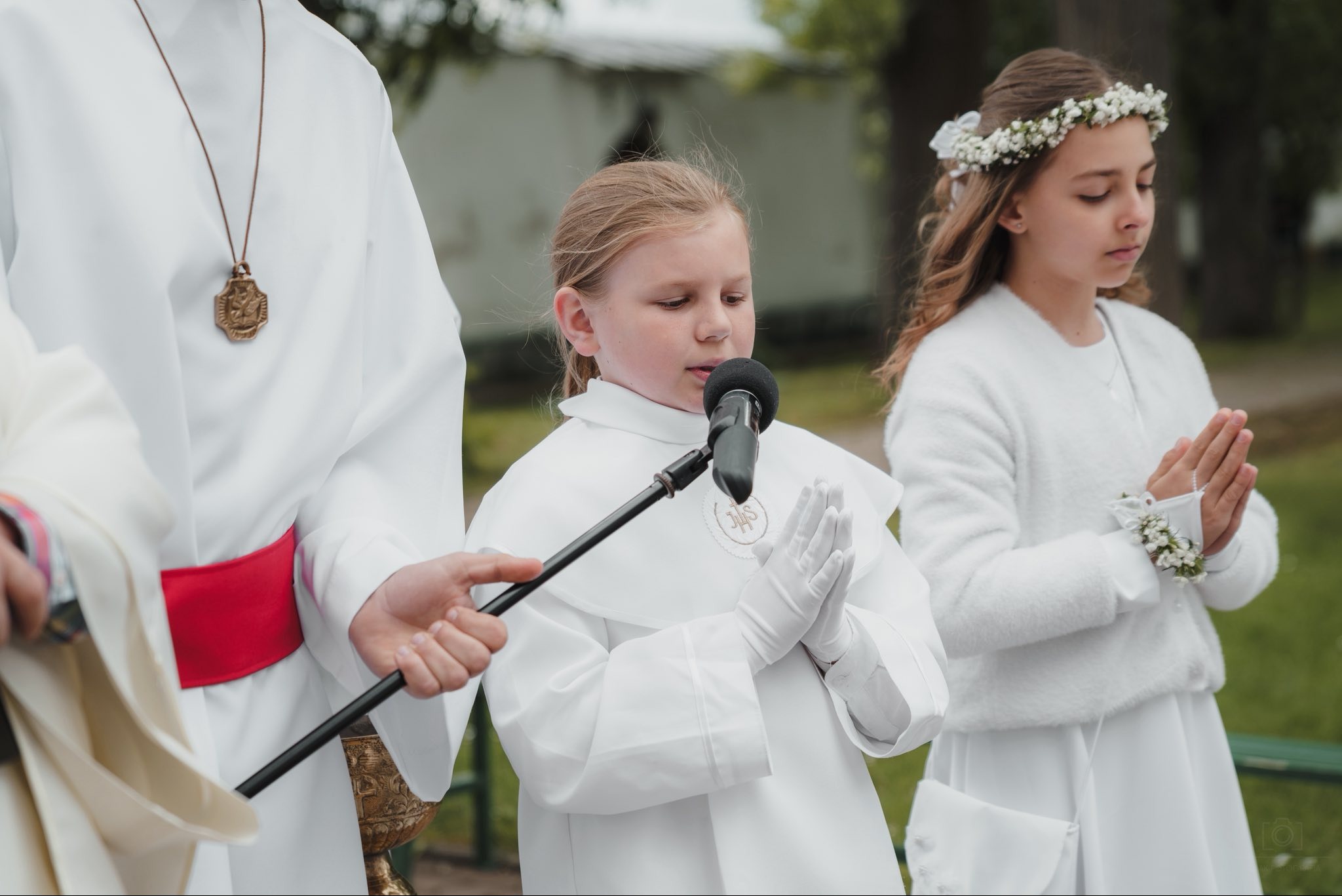 Dziewczynka w albie recytuje tekst do mikrofonu podczas Pierwszej Komunii Świętej w Zakliczynie. W tle druga dziewczynka z wiankiem na głowie i ksiądz.