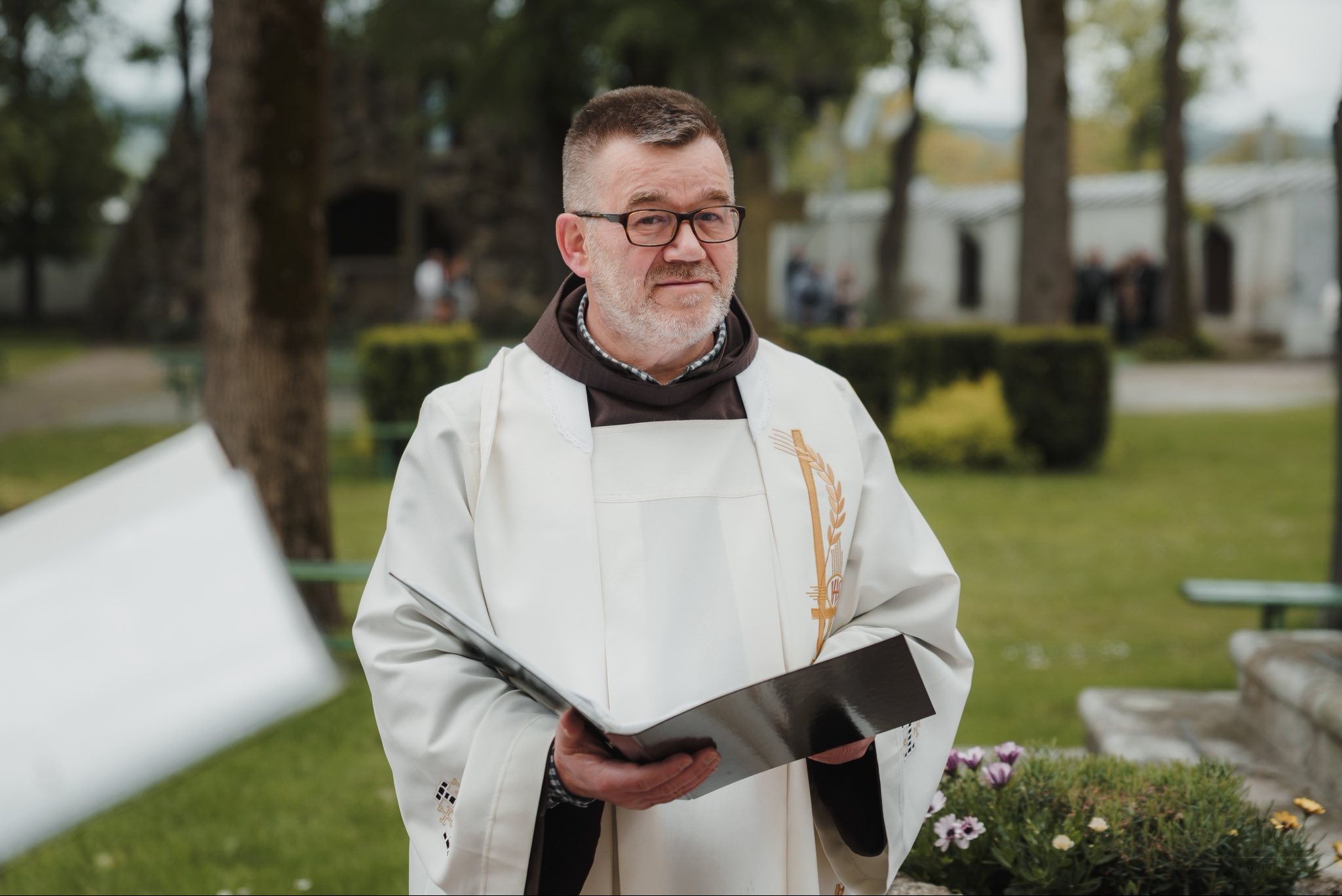 Portret zakonnika w białej szacie z haftem, trzymającego czarny segregator na tle zieleni i budynków. Ujęcie z bliska, skupione na twarzy i stroju.
