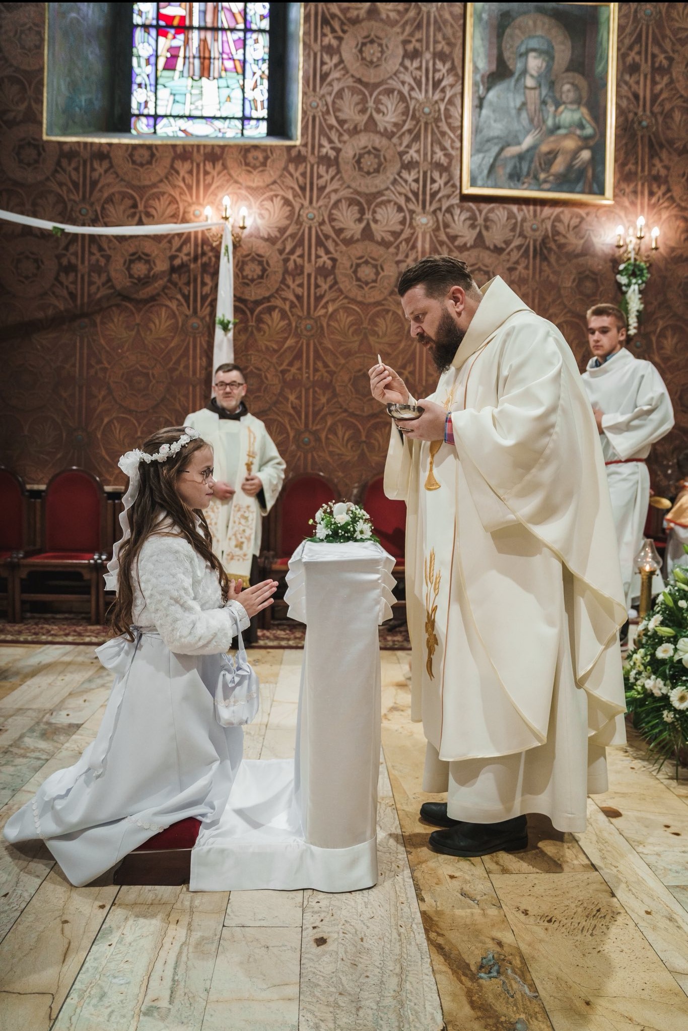 Dziewczynka w białej sukience klęczy przed księdzem podczas ceremonii Pierwszej Komunii Świętej w bogato zdobionym kościele. W tle inni duchowni i witraże.