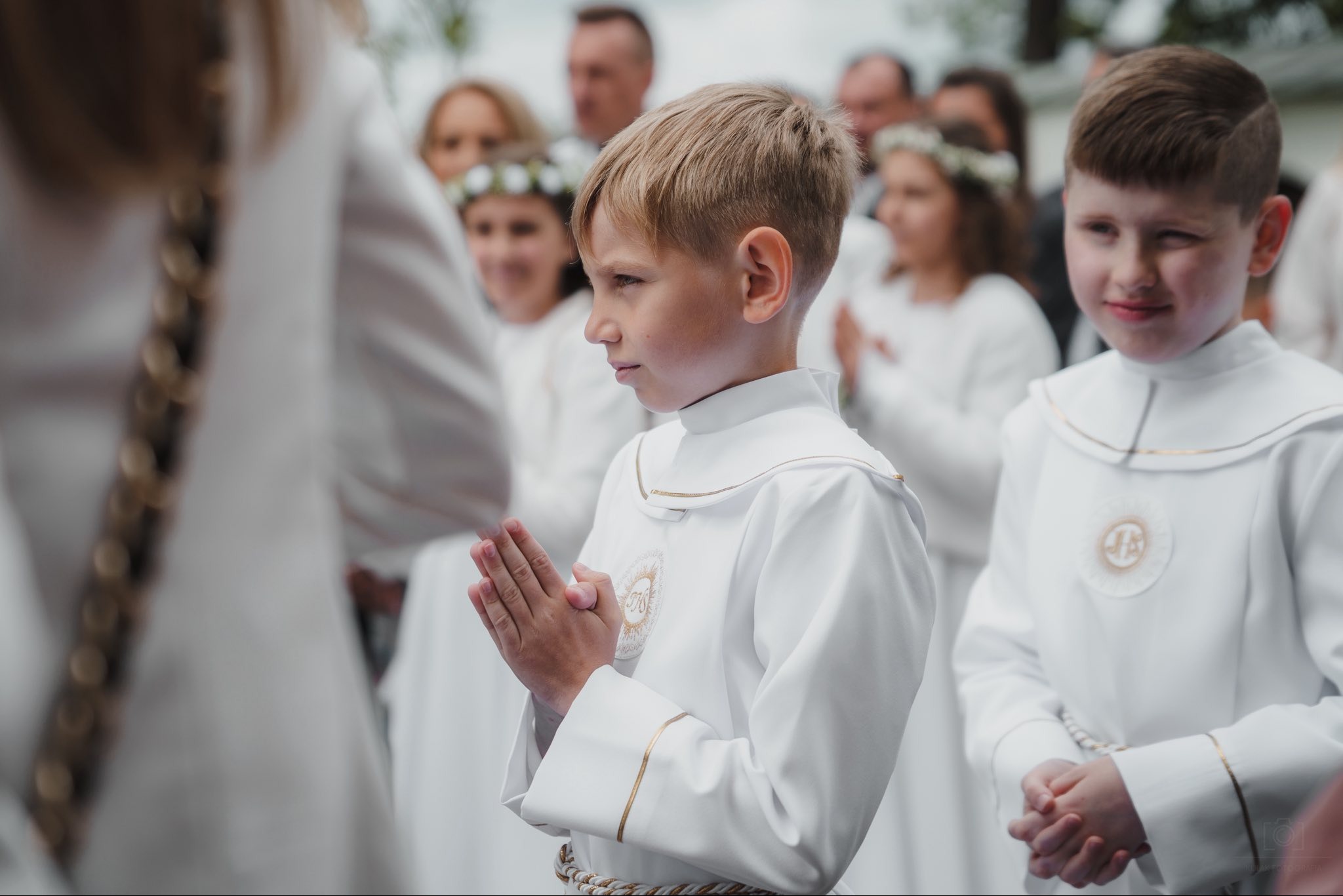 Chłopiec w albie komunijnej, z złączonymi dłońmi, w skupieniu. W tle inne dzieci w białych strojach i dorośli. Ujęcie z delikatnym rozmyciem tła.