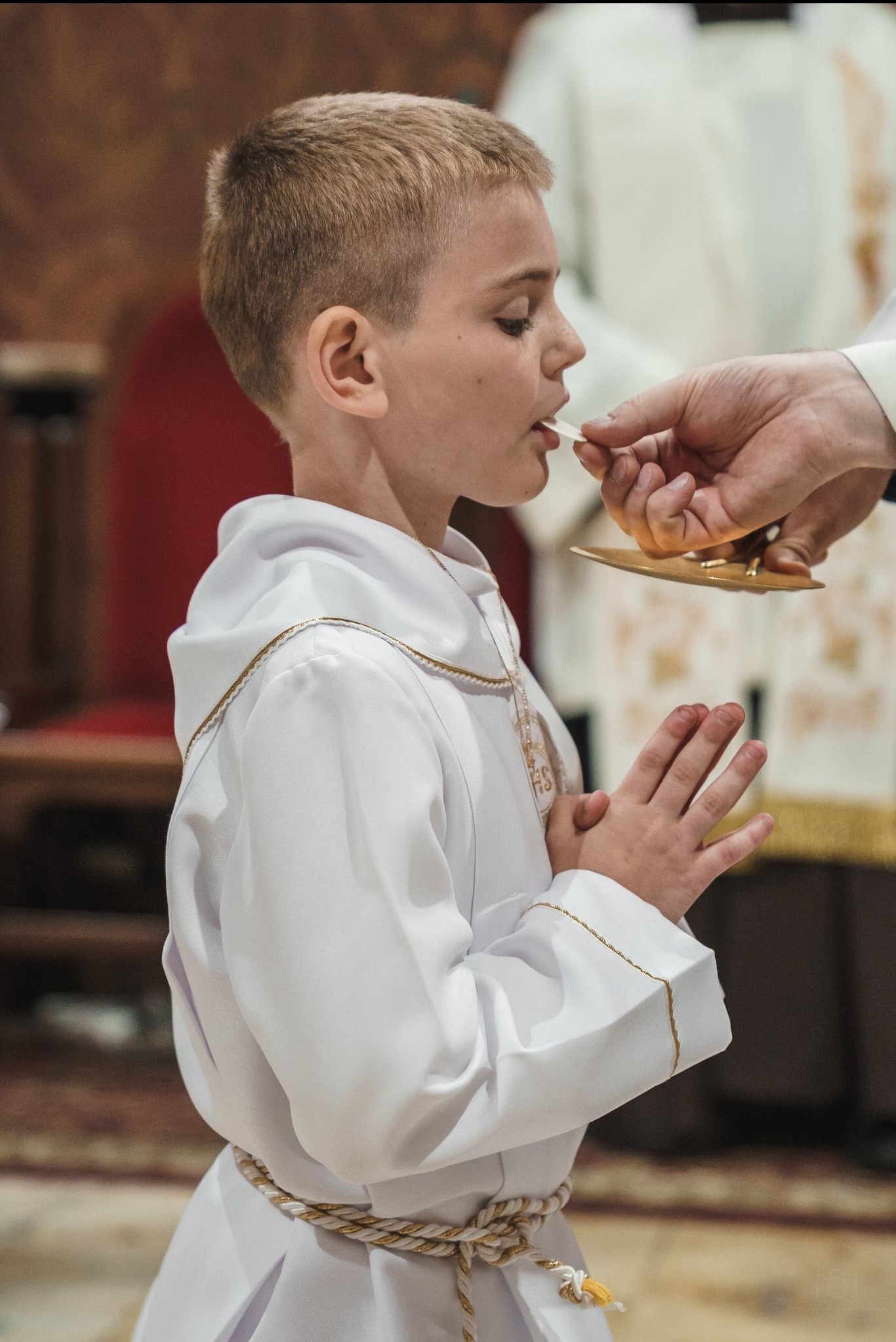 Chłopiec w białej albie przyjmuje komunię świętą, dłonie złożone do modlitwy, delikatne oświetlenie podkreśla uroczysty charakter wydarzenia.