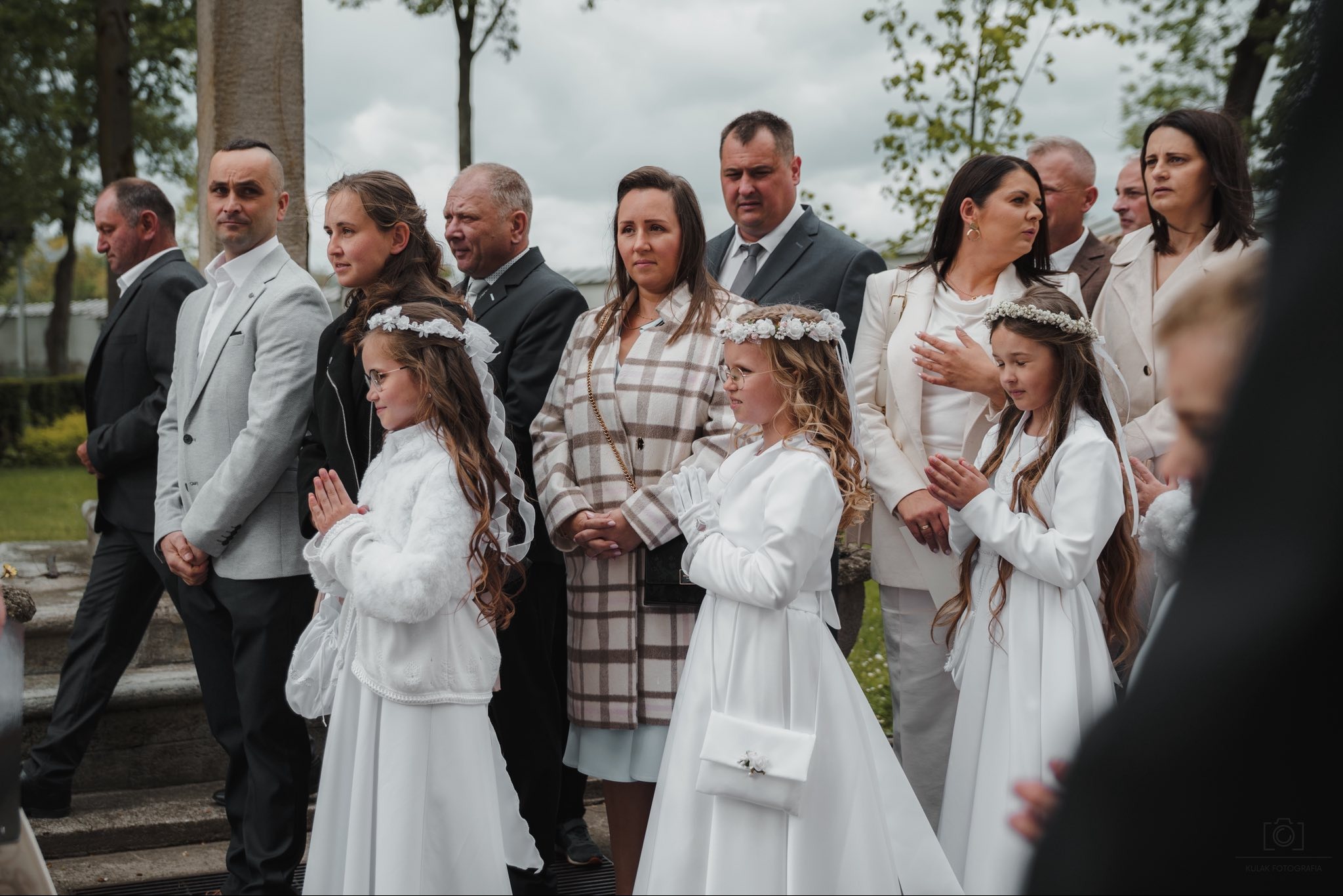 Uroczystość Pierwszej Komunii Świętej: Grupa dzieci w białych albach, z wiankami na głowach, stoi obok rodziny. Ujęcie z zewnątrz, w tle zieleń i fragment kolumny.