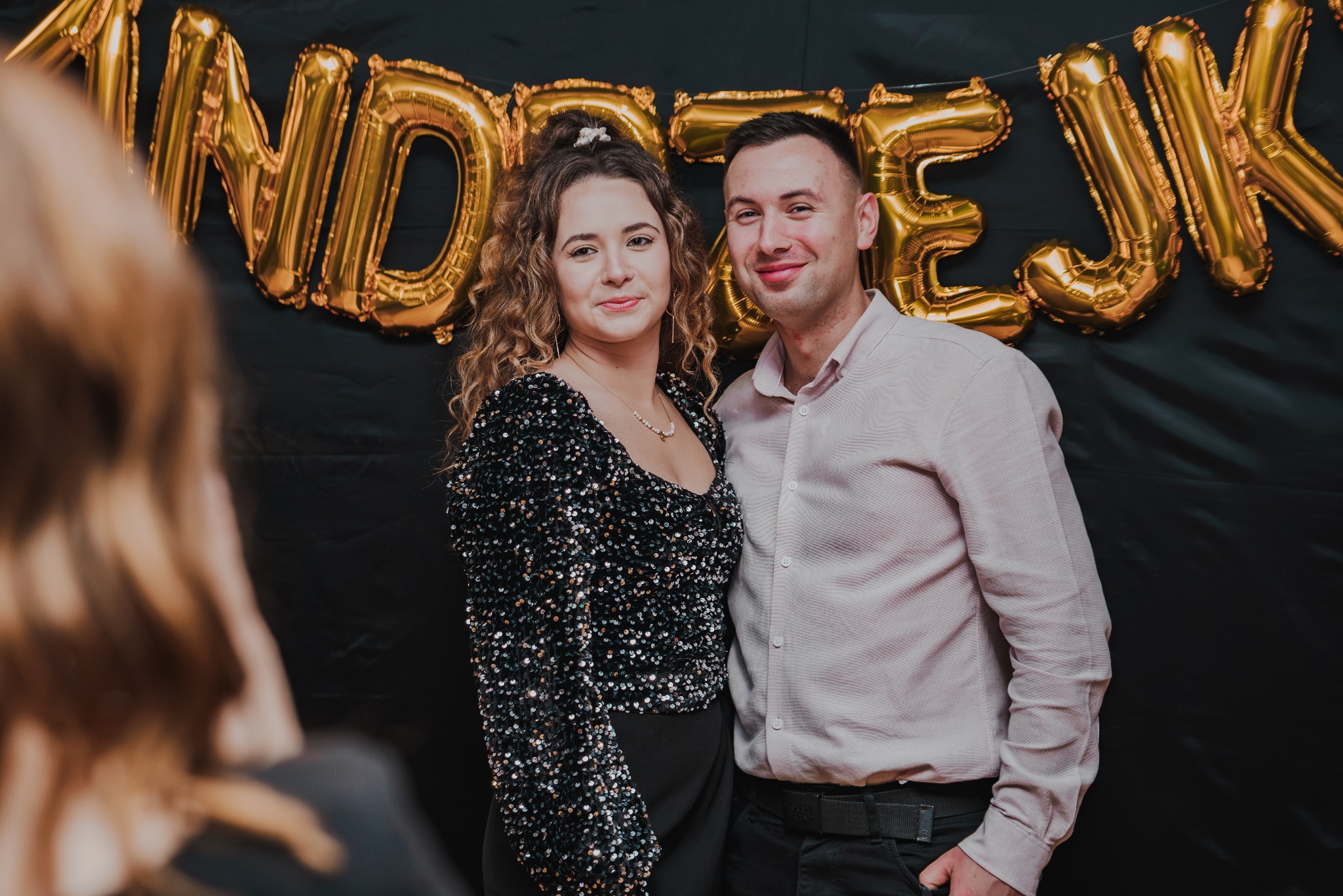 Uśmiechnięta para na tle czarnej ściany z napisem z balonów w kolorze złota. Kobieta w cekinowej bluzce, mężczyzna w koszuli. Ujęcie portretowe.