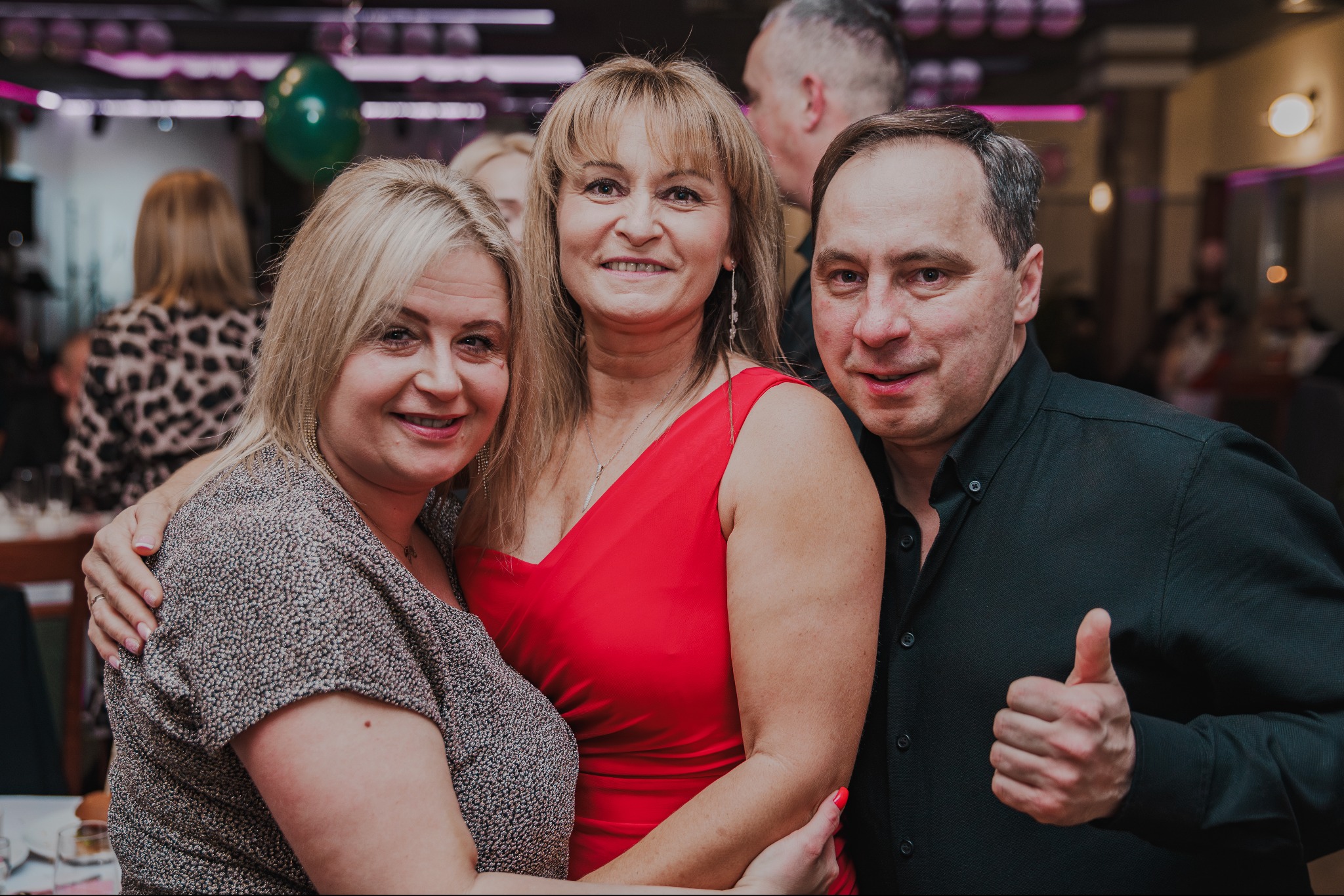 Uśmiechnięta grupa trzech osób pozuje do zdjęcia w pomieszczeniu z dekoracjami. Mężczyzna w czarnej koszuli pokazuje kciuk w górę.