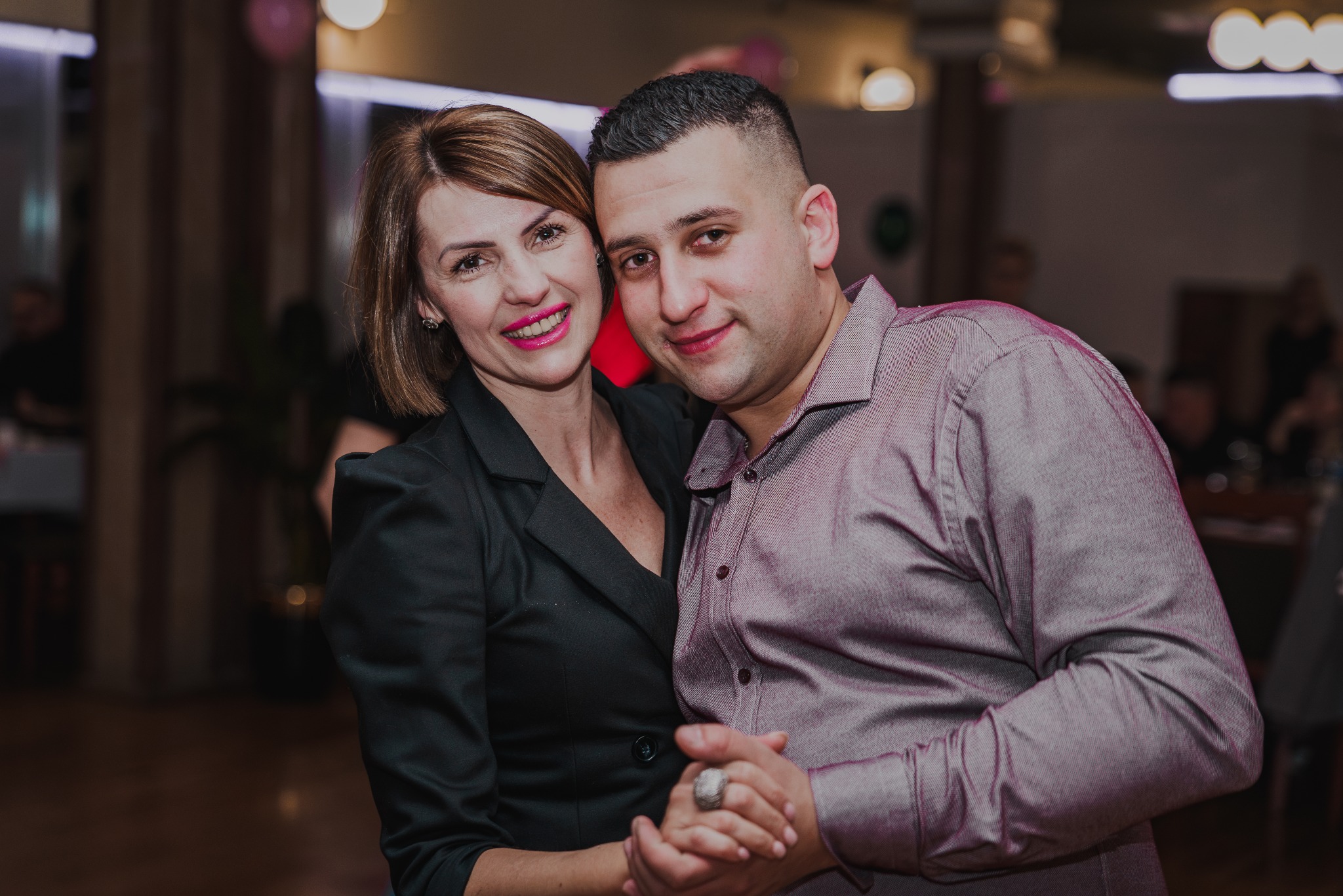 Uśmiechnięta para, kobieta w czarnej marynarce i mężczyzna w bordowej koszuli, pozują blisko siebie na tle sali z dekoracjami.