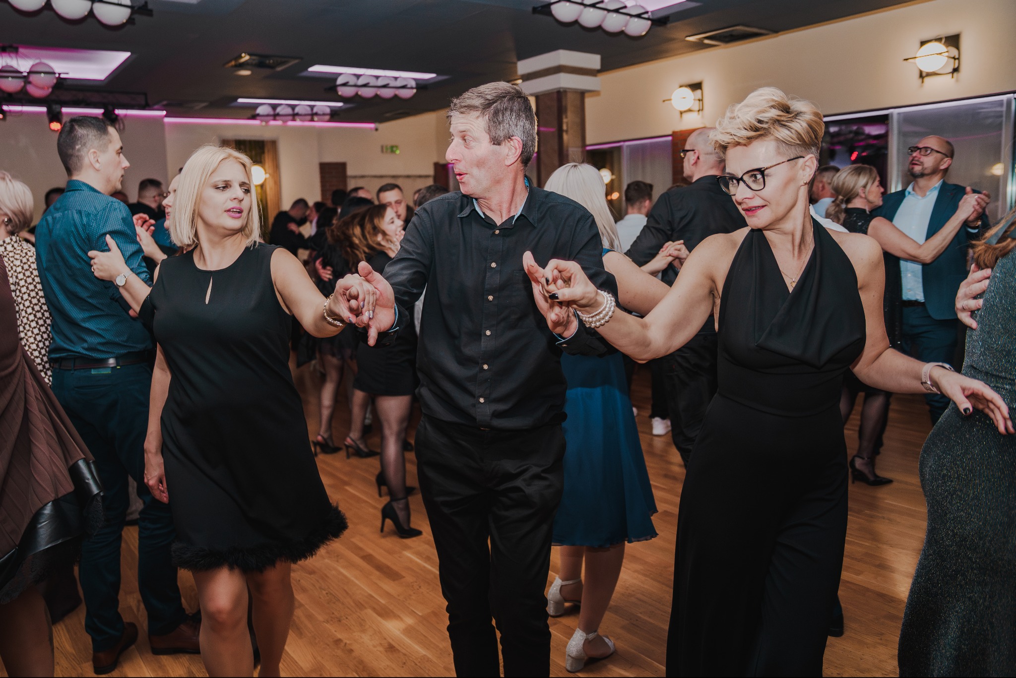 Uczestnicy w trakcie tańca na parkiecie podczas imprezy. W tle widoczni inni goście i elementy dekoracyjne sali.
