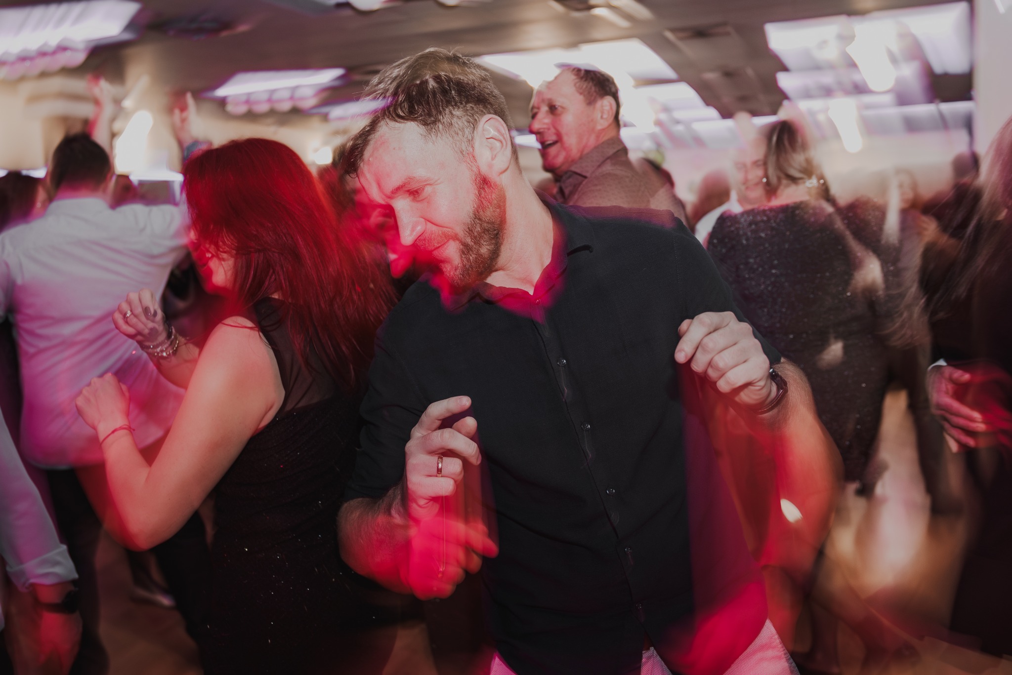 Rozmazane ujęcie z imprezy: mężczyzna w czarnej koszuli tańczy w tłumie, oświetlony czerwonym światłem, z widocznymi innymi osobami w tle. Dynamiczna atmosfera.