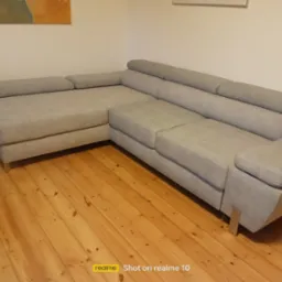 Jasnoszary narożnik z niskim oparciem na drewnianej podłodze. Minimalistyczny design, widoczne metalowe nóżki. Ujęcie z niskiej perspektywy, naturalne oświetlenie.