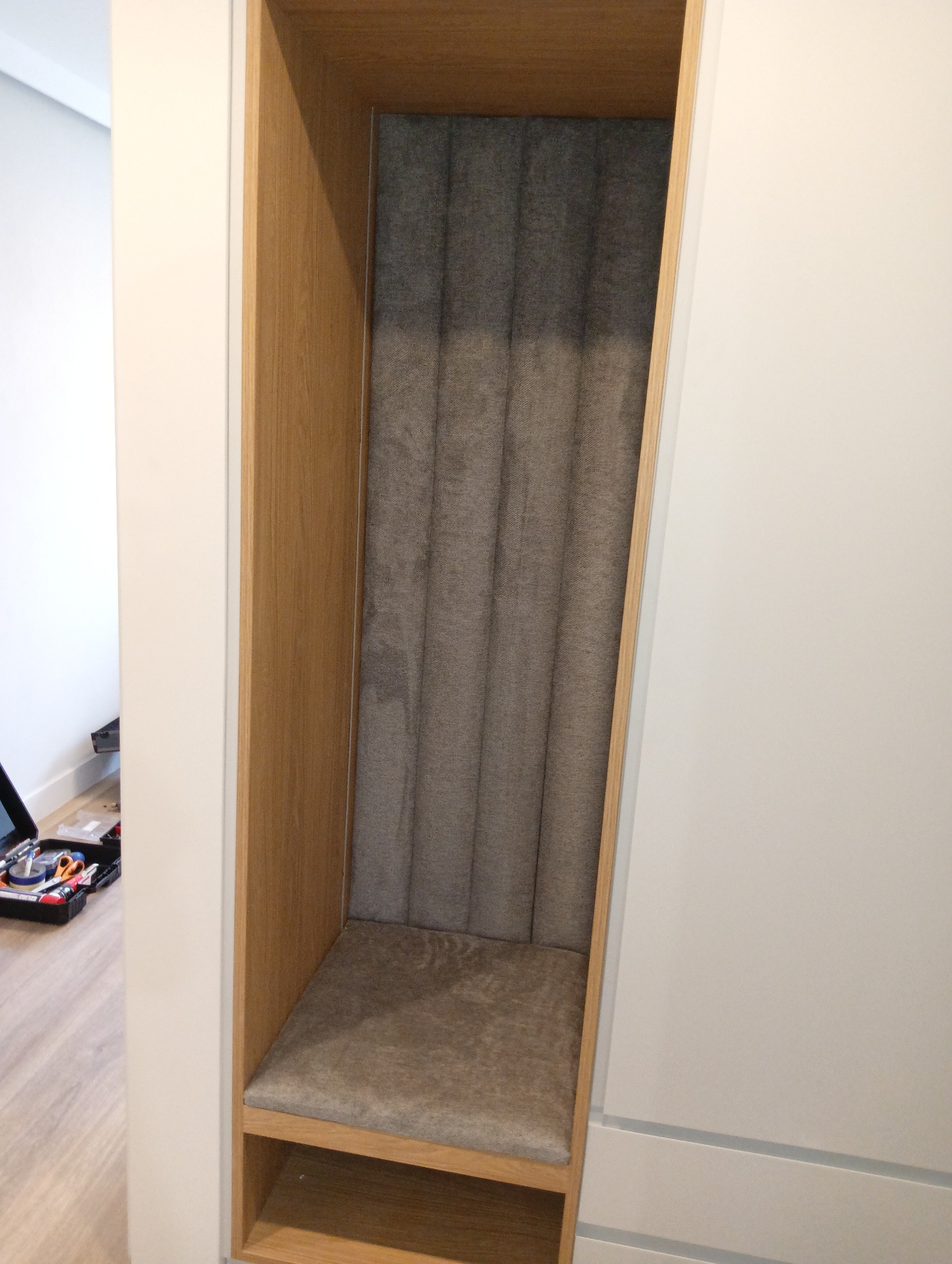 Wnęka z siedziskiem i tapicerowanym panelem ściennym w odcieniach szarości, obudowana drewnem. Widoczny fragment podłogi i narzędzia.