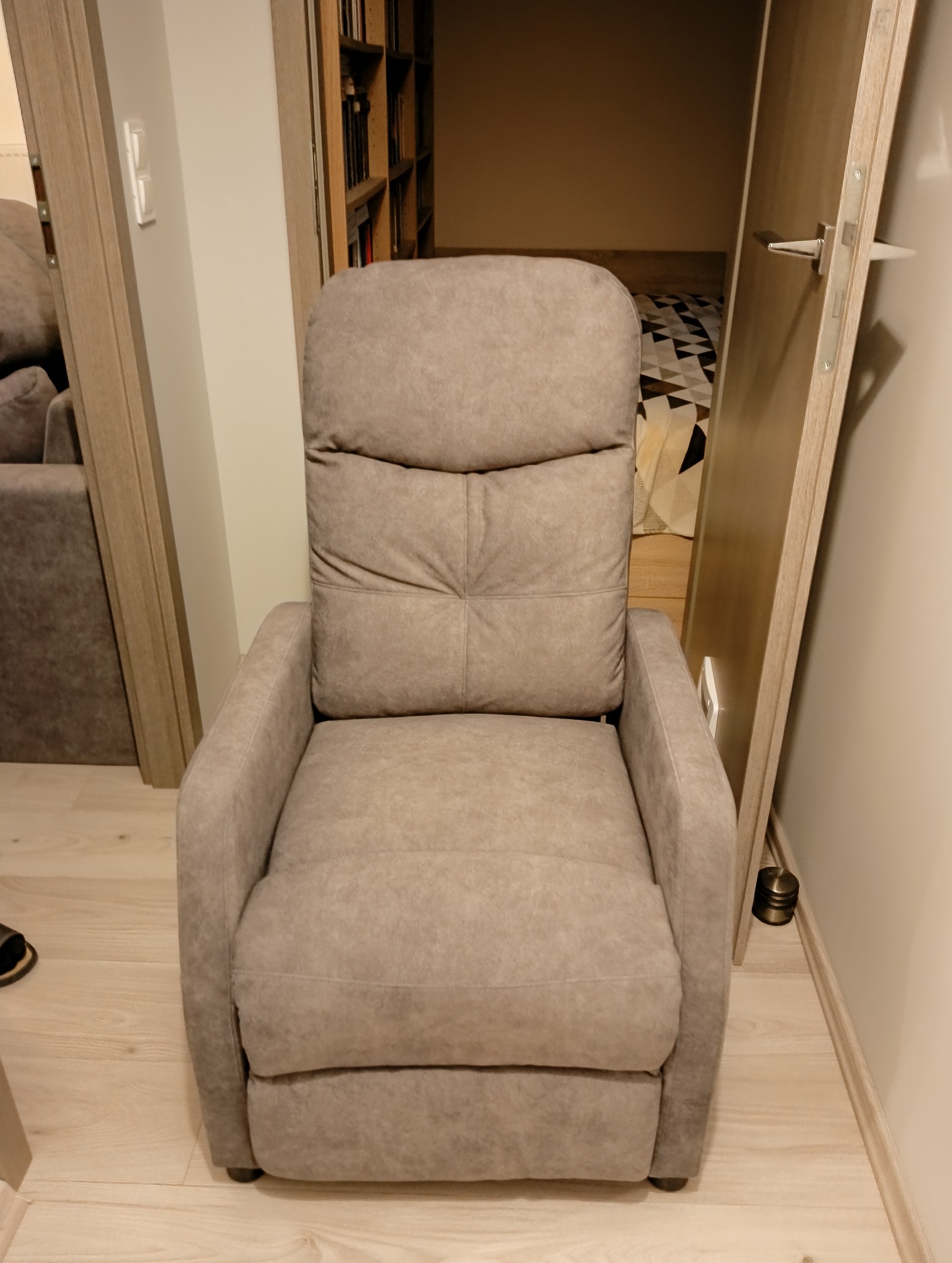 Beżowy, tapicerowany fotel relaksacyjny z wysokim oparciem w pokoju. Widoczny fragment regału z książkami i drzwi. Fotel na drewnianej podłodze.