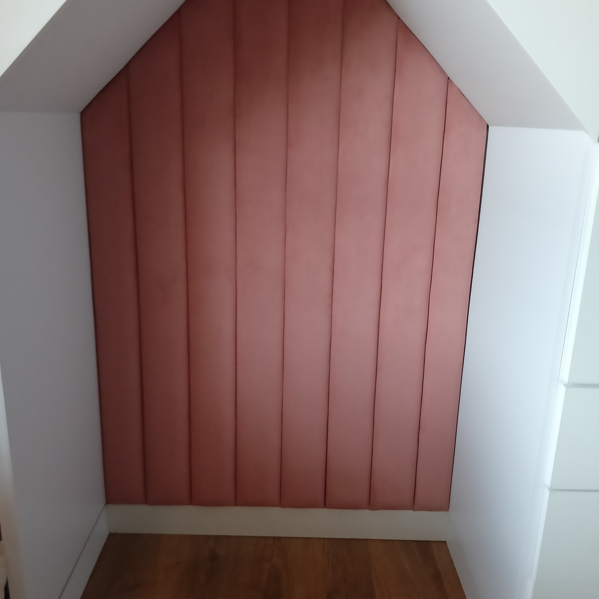 Tapicerowane panele ścienne w odcieniach różu, dopasowane do skosu poddasza. Widoczna drewniana podłoga i białe elementy zabudowy.