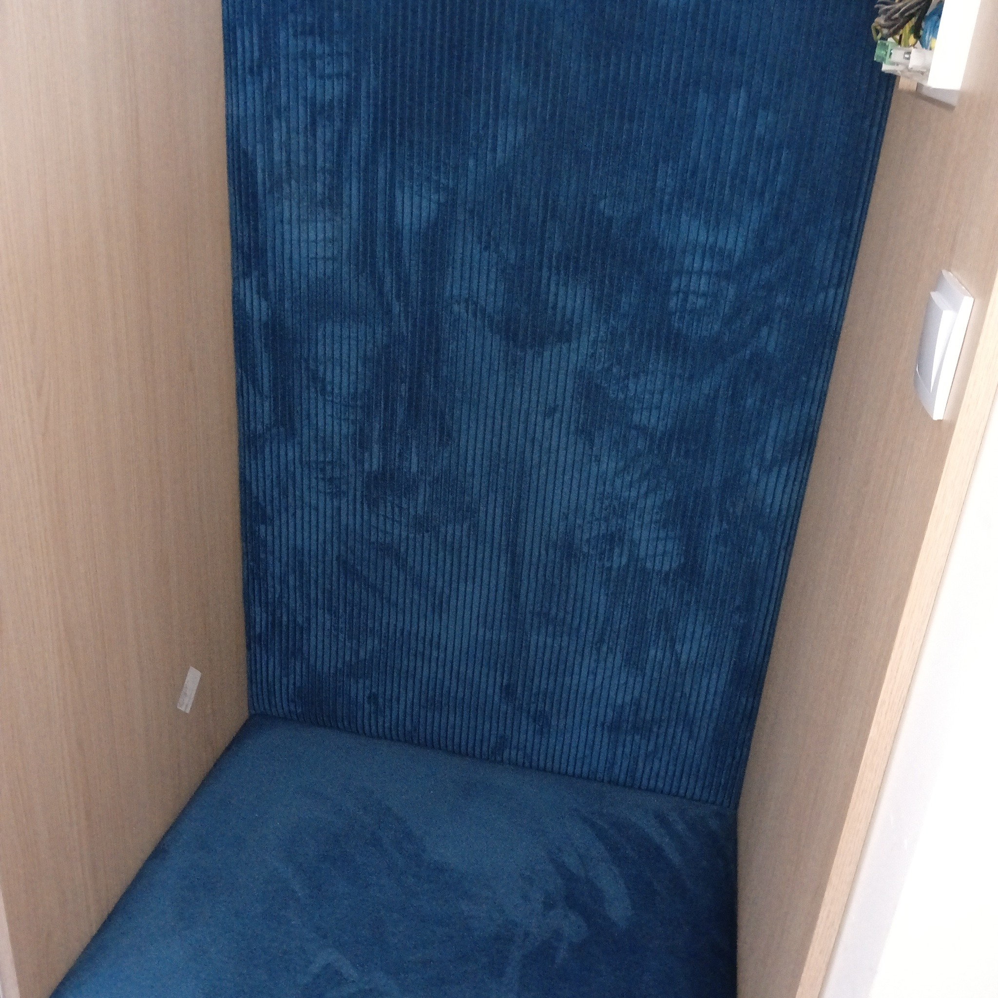 Narożnik z niebieską tapicerką w drewnianej wnęce. Pionowe prążki materiału, jasne drewno, minimalistyczny design. Tapicerowane siedzisko i oparcie.