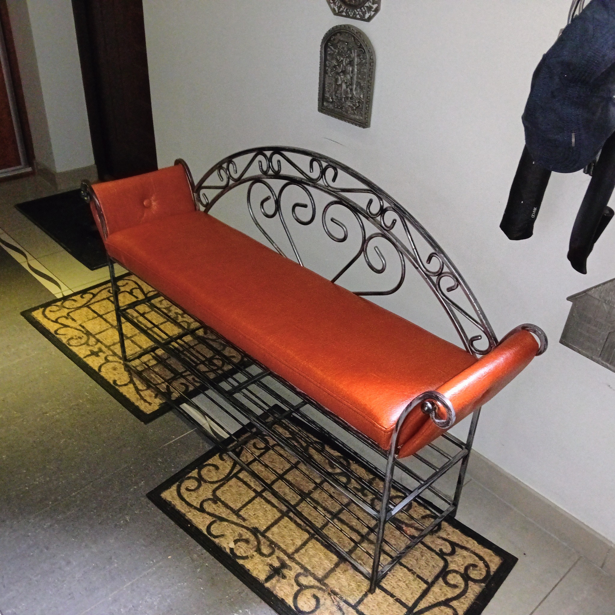 Metalowa ławka z pomarańczowym siedziskiem na tle ściany z dekoracjami. Ławka stoi na wycieraczkach z wzorem. Widok z góry, oświetlenie dzienne.