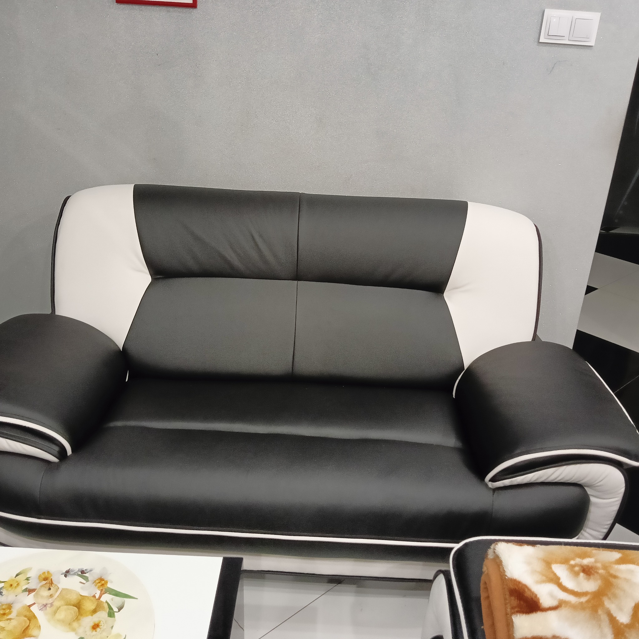 Dwukolorowa, skórzana sofa (czarno-biała) z widocznymi detalami przeszyć i podłokietników. Fragment stolika kawowego z dekoracją kwiatową w kadrze.