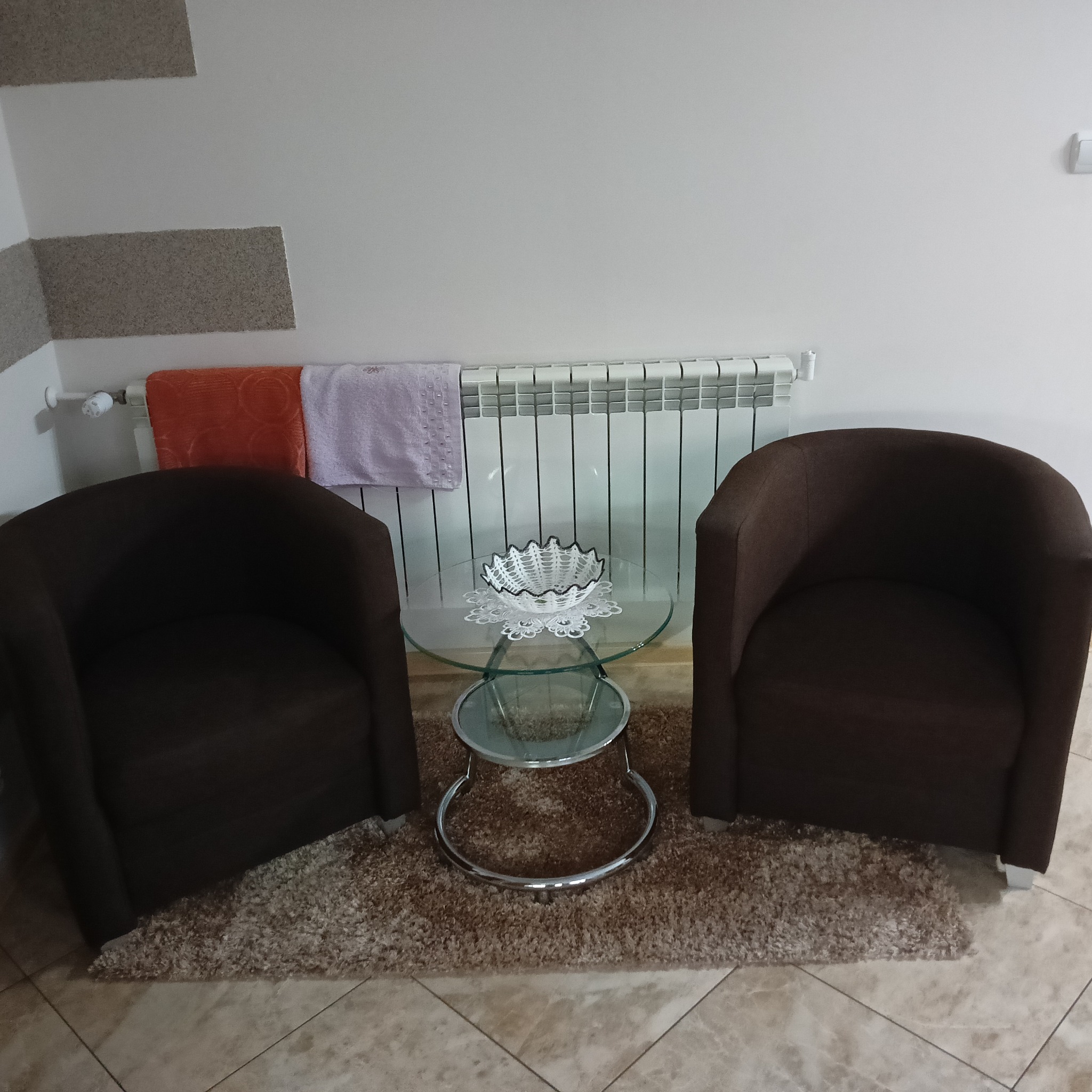 Dwa brązowe fotele klubowe z niskim, okrągłym stolikiem ze szklanym blatem na dywanie typu shaggy. Na grzejniku wiszą ręczniki.