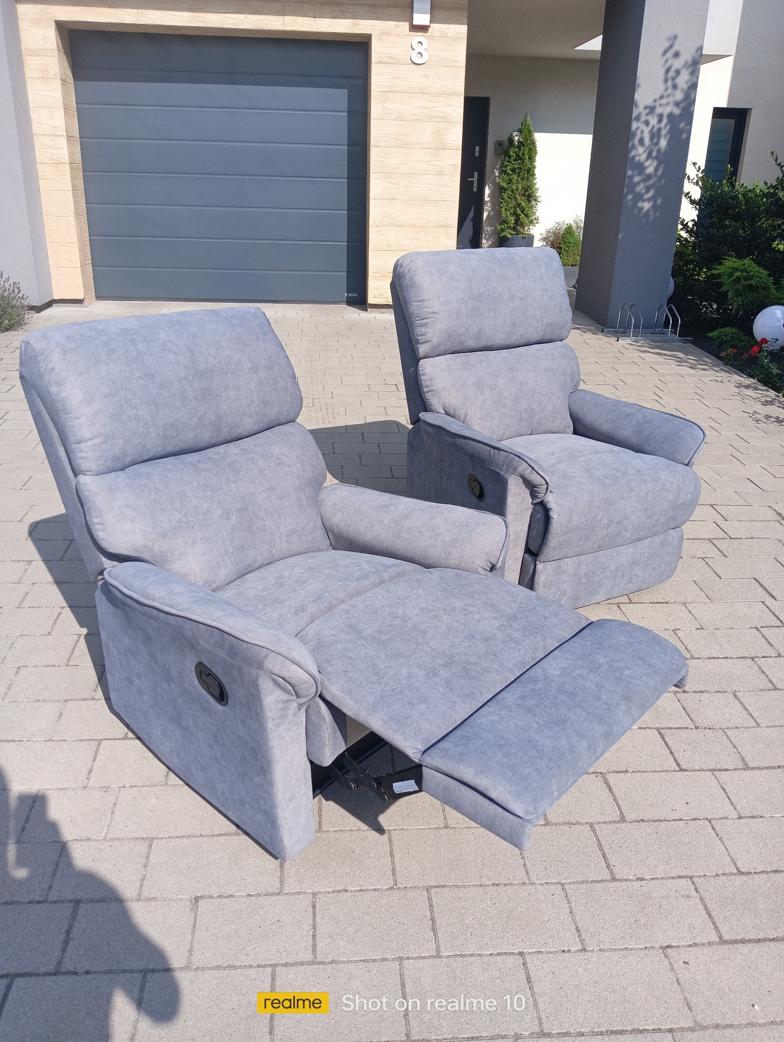Dwa szare fotele tapicerowane z wysuwanymi podnóżkami na tle nowoczesnego domu z garażem i zielenią. Jeden fotel z podnóżkiem wysuniętym, drugi złożony.