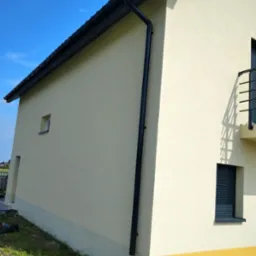 Jasna elewacja domu z czarną rynną i oknami z żaluzjami. Dół ściany w kolorze żółtym, kontrastujący z resztą elewacji. Błękitne niebo w tle.