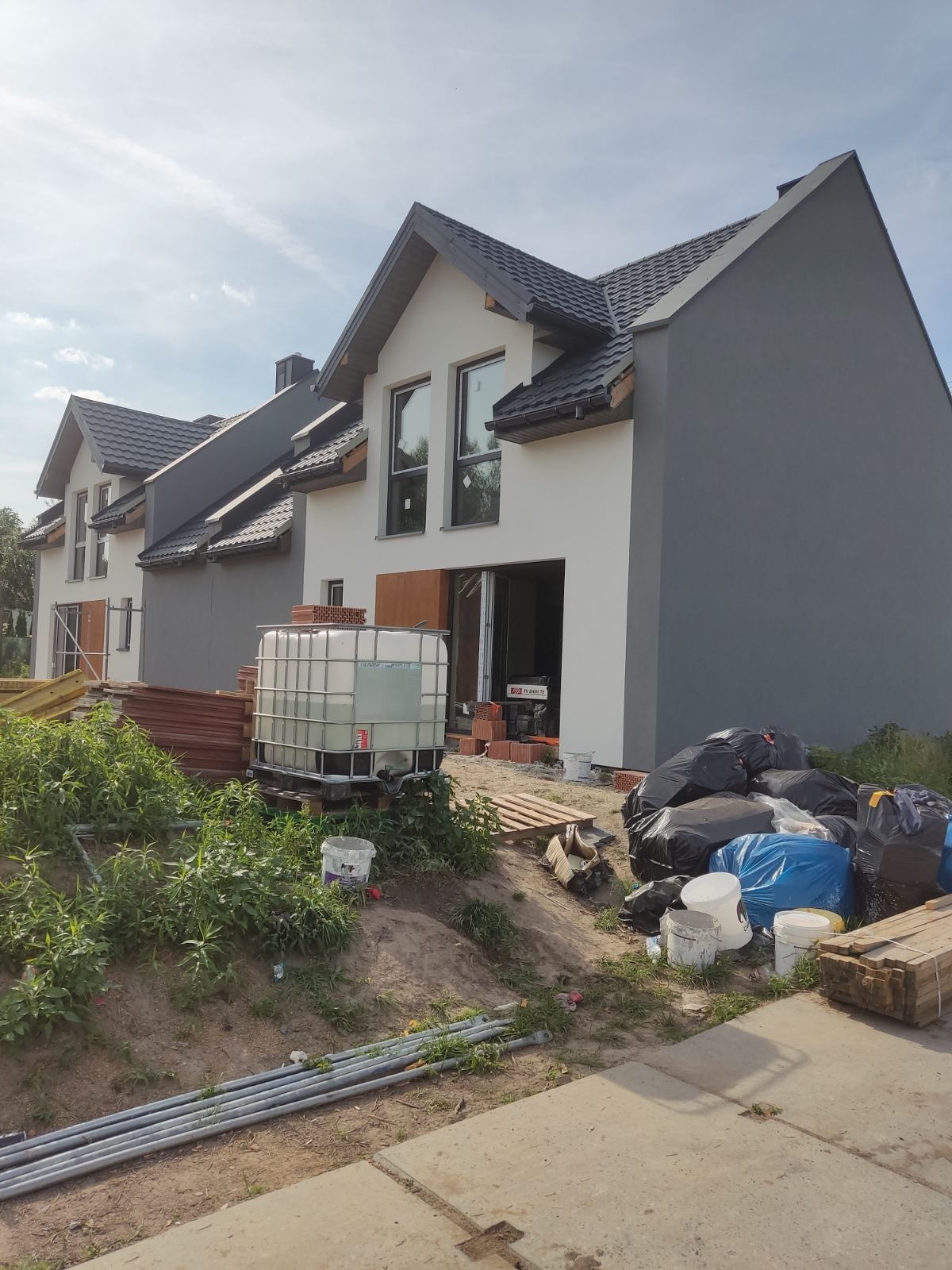 Domy w trakcie budowy z częściowo pomalowaną elewacją na biało i szaro, widoczne materiały budowlane i zbiornik na wodę. Prace w toku.