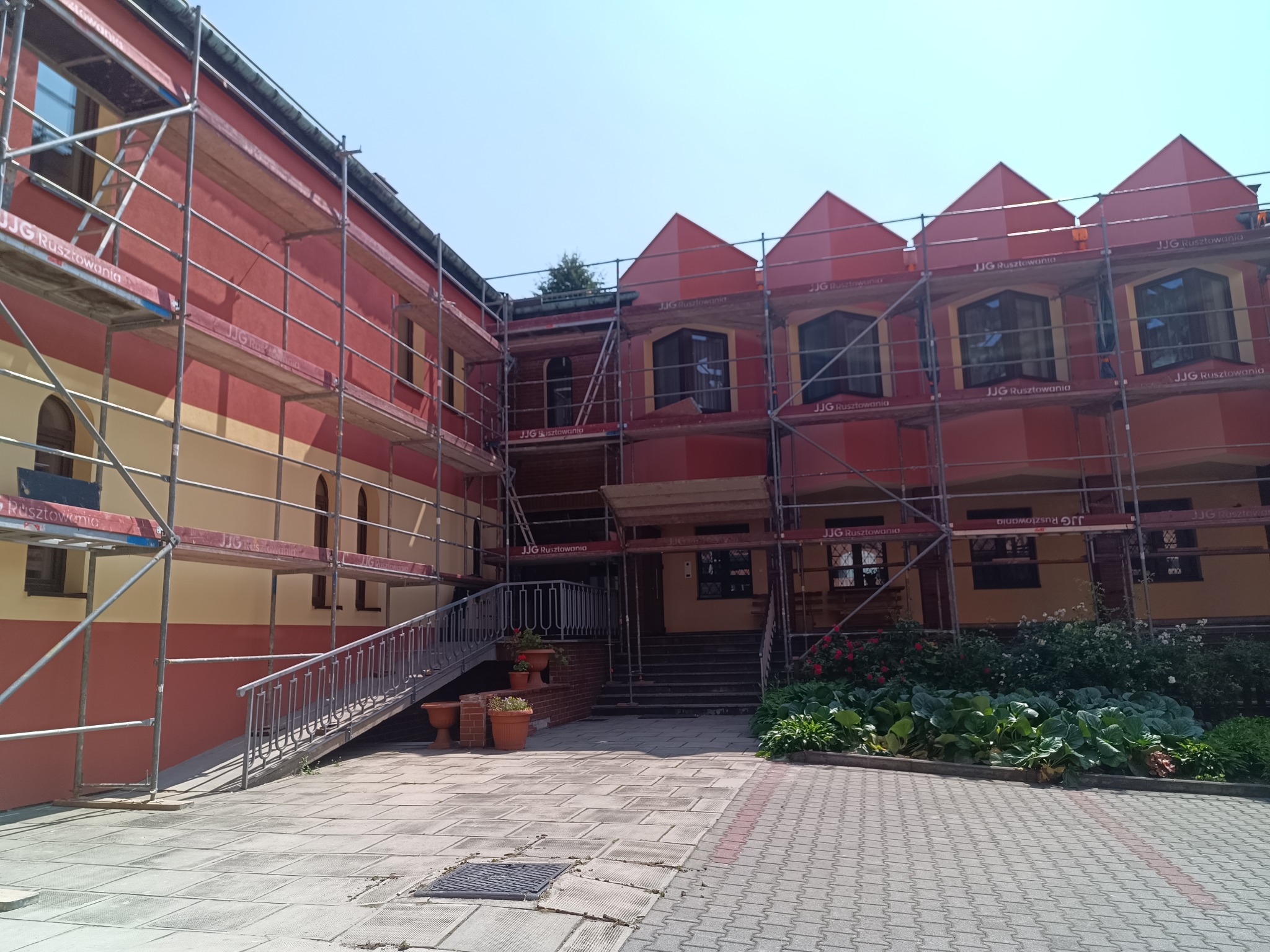 Budynek w trakcie prac elewacyjnych, otoczony rusztowaniami. Widoczne fragmenty pomalowanej fasady w kolorach czerwonym i żółtym. Na pierwszym planie chodnik z kostki.