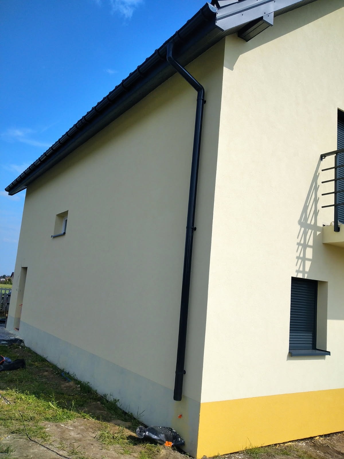 Jasna elewacja domu z czarną rynną i oknami z żaluzjami. Dół ściany w kolorze żółtym, kontrastujący z resztą elewacji. Błękitne niebo w tle.