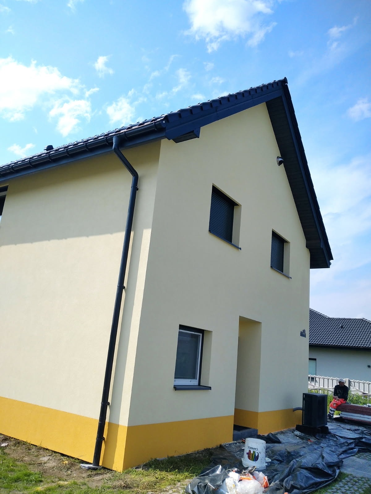 Otynkowana elewacja domu jednorodzinnego z fragmentem pomalowanej na żółto dolnej części. Widoczne okna z roletami, rynna i fragment nieba z chmurami.