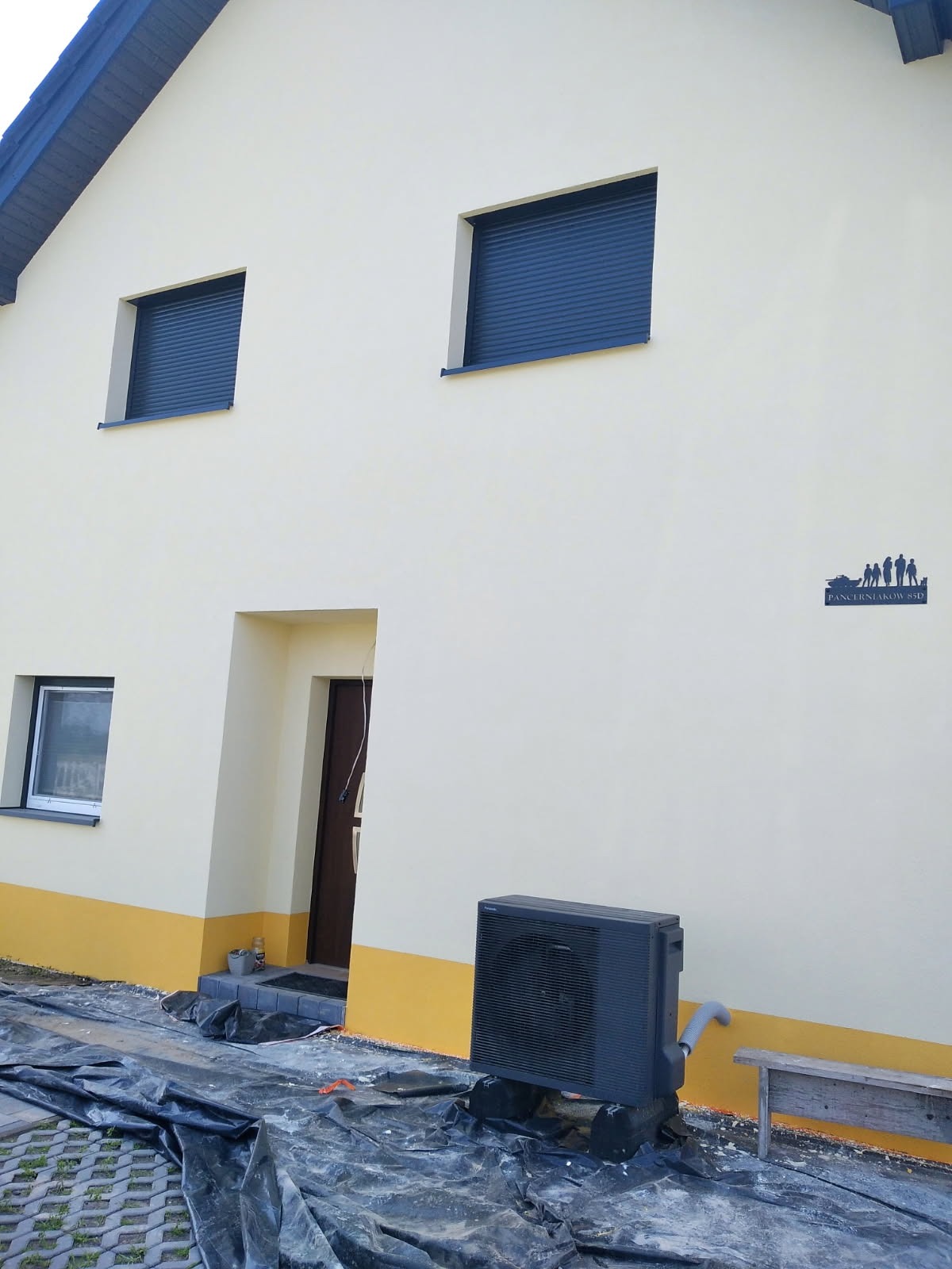 Jasna elewacja domu z niebieskimi roletami i dekoracją ścienną. Widoczny fragment żółtej, dolnej części elewacji i czarna jednostka zewnętrzna klimatyzacji.
