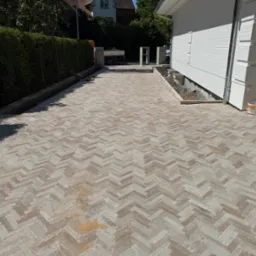 Nowo ułożona kostka brukowa w jodełkę na podjeździe, z widocznymi obrzeżami i elementami wykończeniowymi w pobliżu białego budynku. Słoneczny dzień.