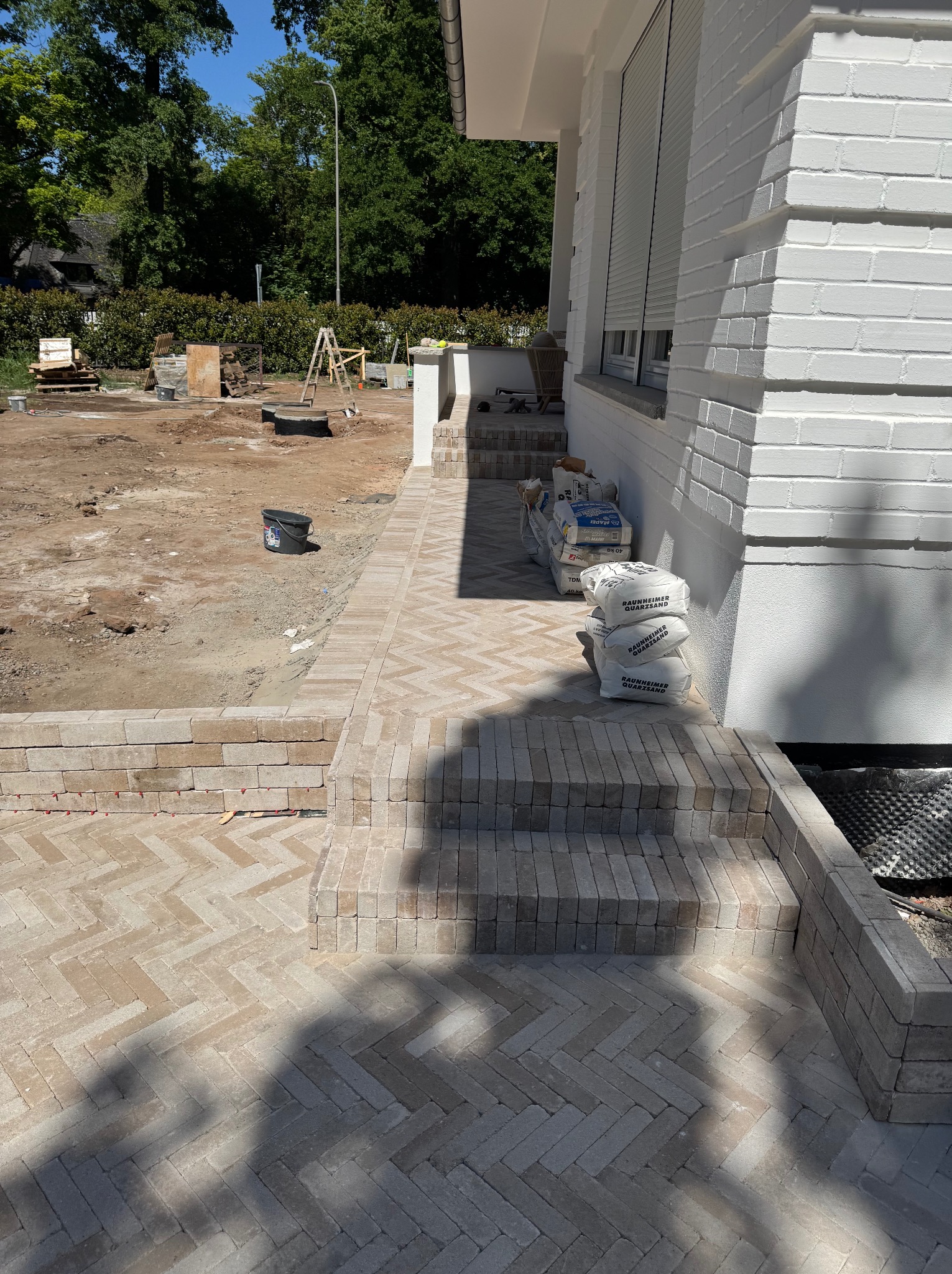 Nowa, beżowa kostka brukowa ułożona w jodełkę, tworząca ścieżkę i schody przy białym budynku. Worki z zaprawą w tle, teren przygotowany do dalszych prac.