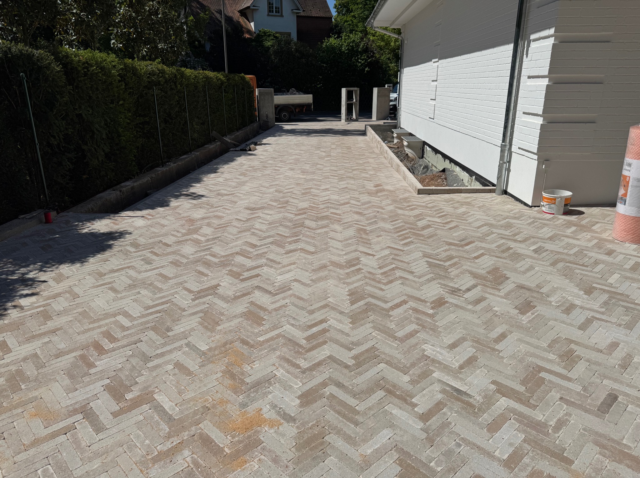 Nowo ułożona kostka brukowa w jodełkę na podjeździe, z widocznymi obrzeżami i elementami wykończeniowymi w pobliżu białego budynku. Słoneczny dzień.