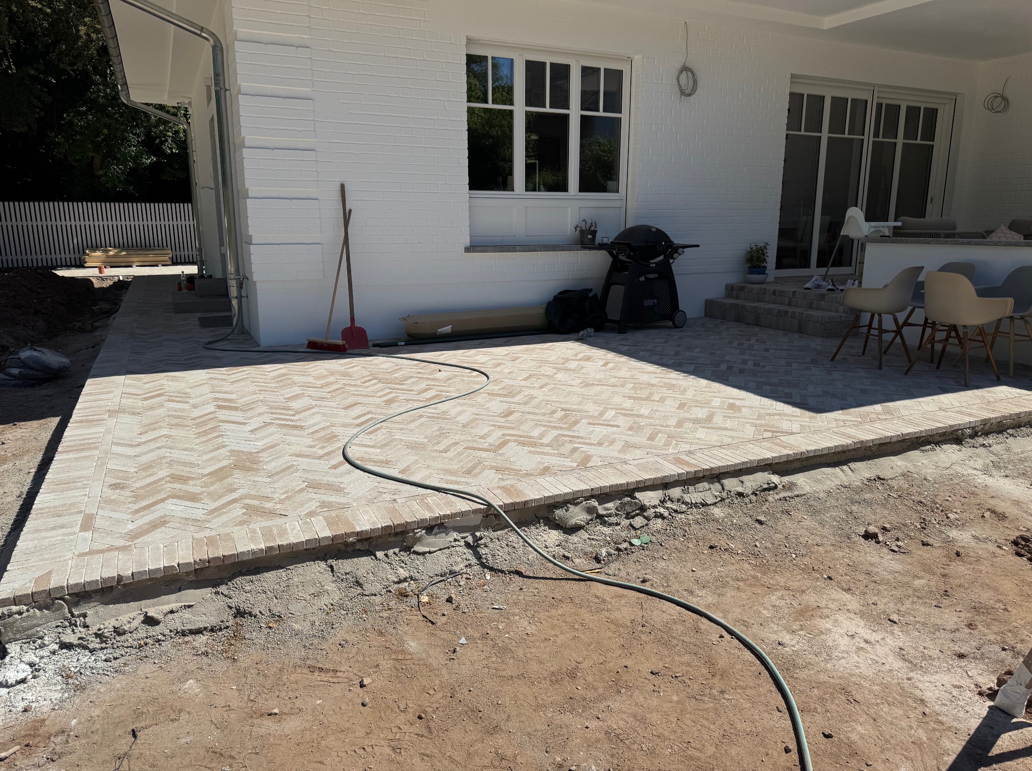 Nowo układana kostka brukowa w jodełkę na tarasie, widoczny fragment posesji z białym budynkiem w tle i narzędzia. Prace w toku, teren przygotowany pod dalsze etapy.