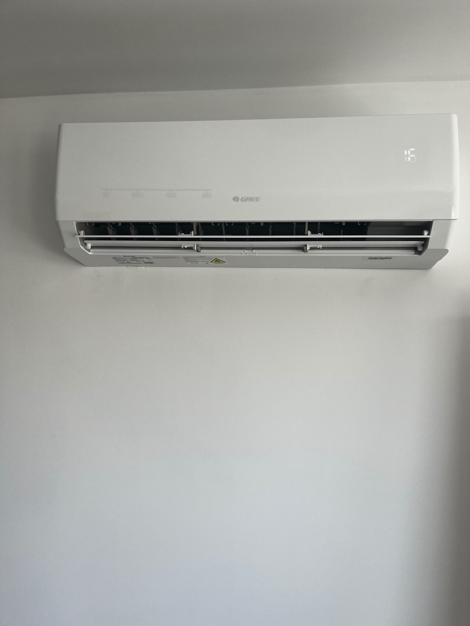 Świeżo zamontowany, biały klimatyzator marki Gree na jasnej ścianie. Widoczny wyświetlacz temperatury i otwarte żaluzje wylotu powietrza. Minimalistyczny design.