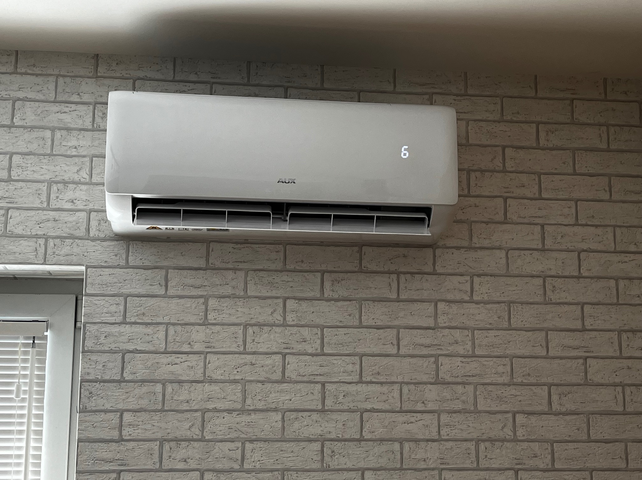 Biały klimatyzator marki AUX zamontowany na ścianie z imitacji cegły. Widoczny wyświetlacz z cyfrą 6. Zamknięty nawiew. Fragment okna z żaluzjami po lewej.