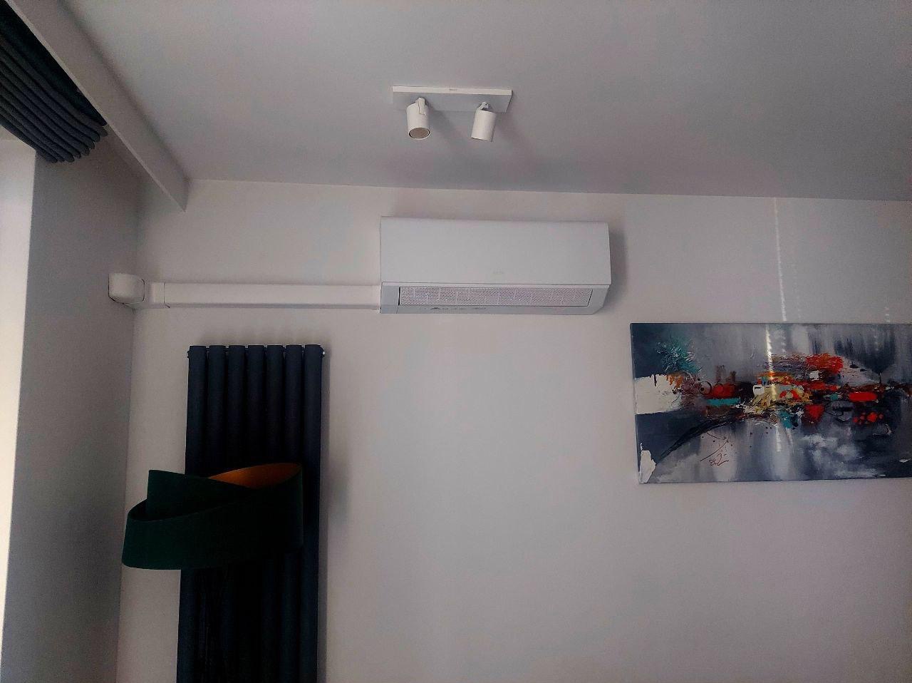 Biała klimatyzacja ścienna w nowoczesnym wnętrzu z grzejnikiem żeberkowym i abstrakcyjnym obrazem. Minimalistyczny design, oświetlenie punktowe.