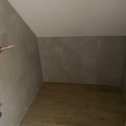 VIP Transport Grzegorz Kożuszkiewicz - Ukończona łazienka na poddaszu z szarymi płytkami ściennymi i drewnopodobną podłogą. Uwagę przyciąga nietypowy kształt pomieszczenia.