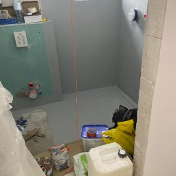 VIP Transport Grzegorz Kożuszkiewicz - Przygotowana do dalszych prac łazienka: szare ściany, uszczelniona podłoga, instalacja wodna ukryta za płytą gipsową. Widoczne materiały budowlane i narzędzia. Okno VEKA w tle.