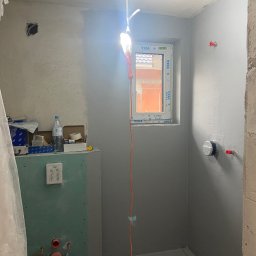 VIP Transport Grzegorz Kożuszkiewicz - Łazienka w trakcie prac wykończeniowych: szpachlowane ściany, zabudowa z płyty GK, okno z widokiem na ceglany budynek, wisząca lampa robocza.