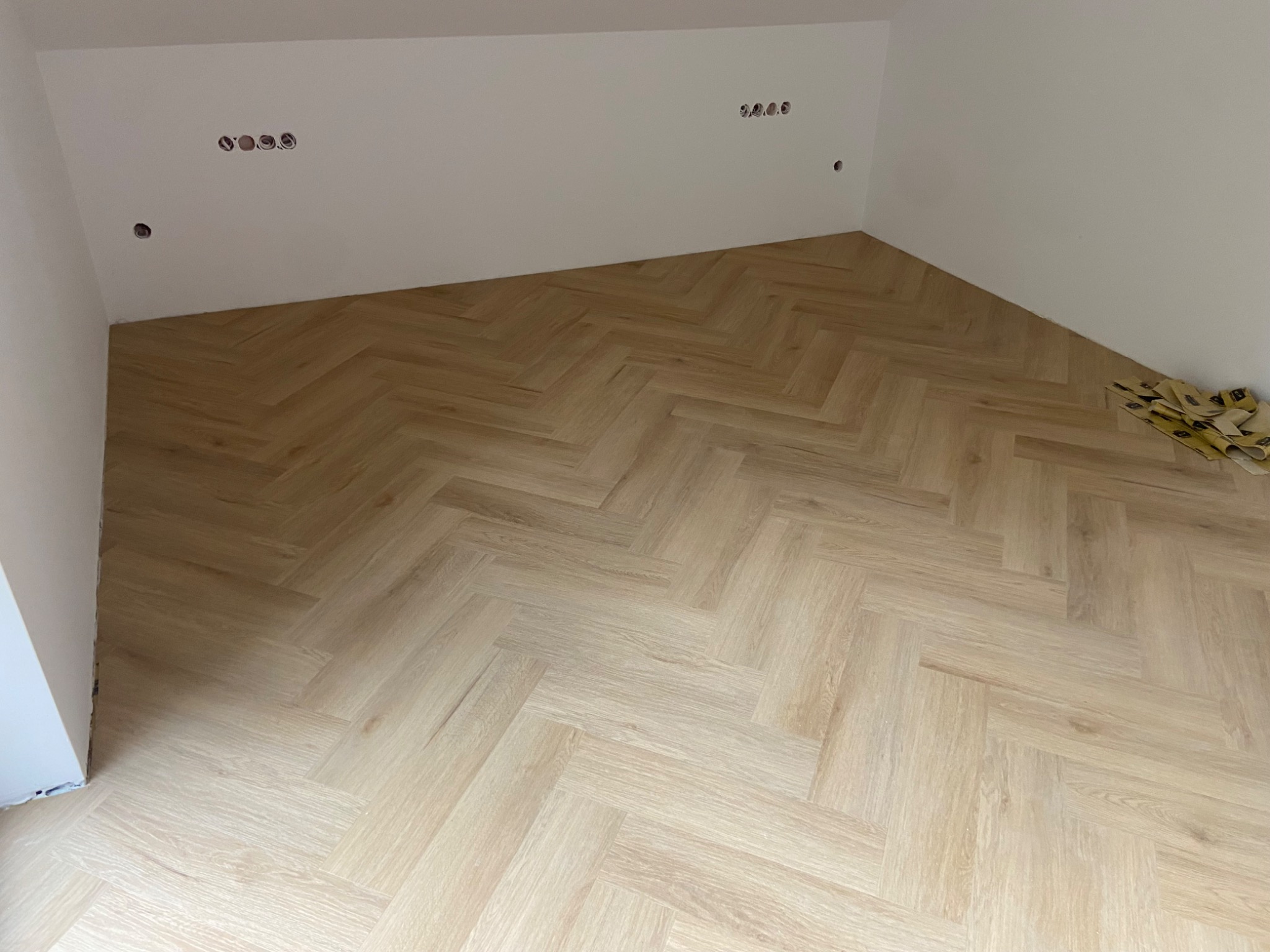 Jasny parkiet ułożony w jodełkę w rogu pokoju z białymi ścianami i widocznymi puszkami elektrycznymi. Na podłodze leżą resztki opakowań po produkcie.