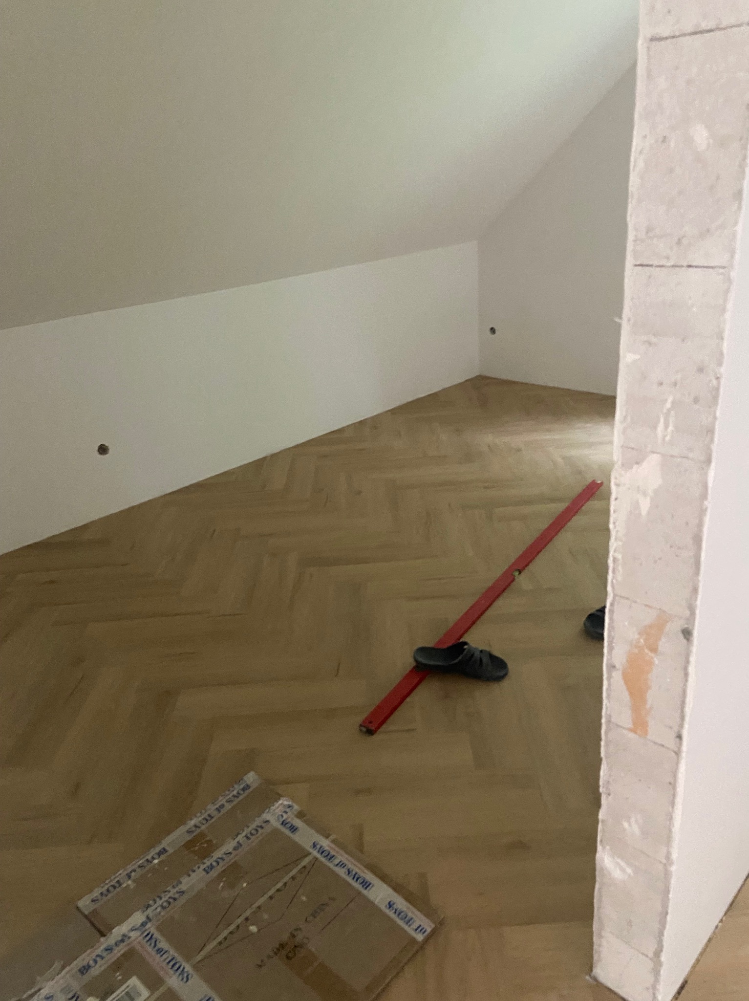 Układanie parkietu w jodełkę w skośnym pokoju. Widoczny poziomica, pudełka po parkiecie i fragment ściany z surowego betonu. Podłoga w trakcie układania.