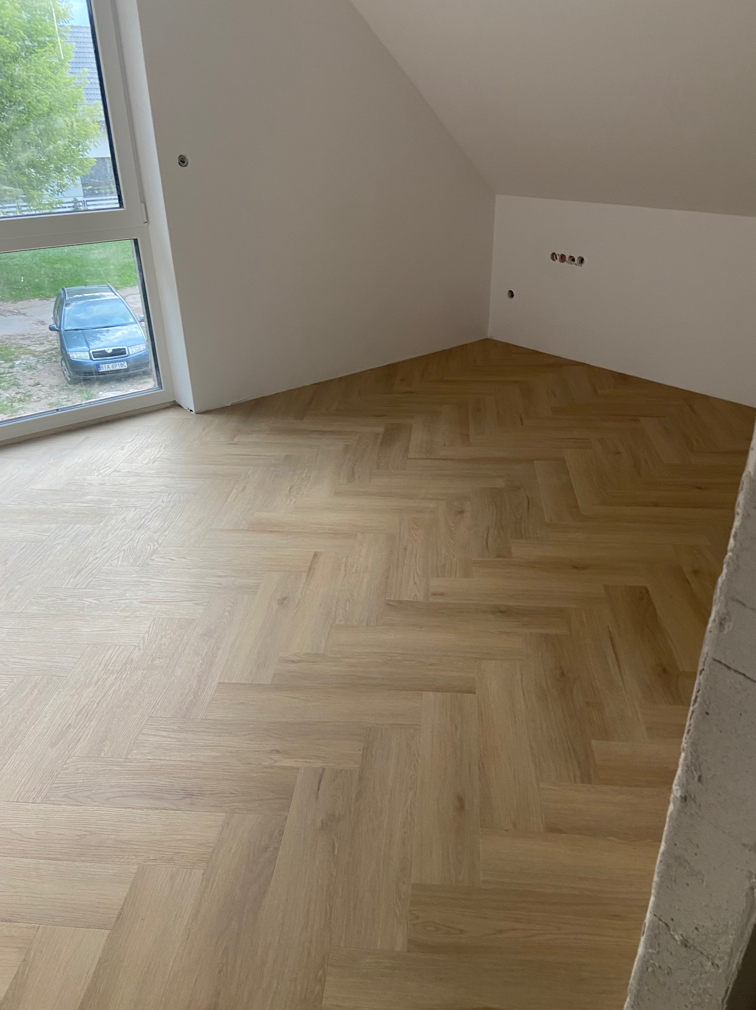 Jasny parkiet ułożony we wzór jodełki francuskiej w nowym pokoju na poddaszu z oknem. Widoczne gniazdka elektryczne i fragment surowej ściany.