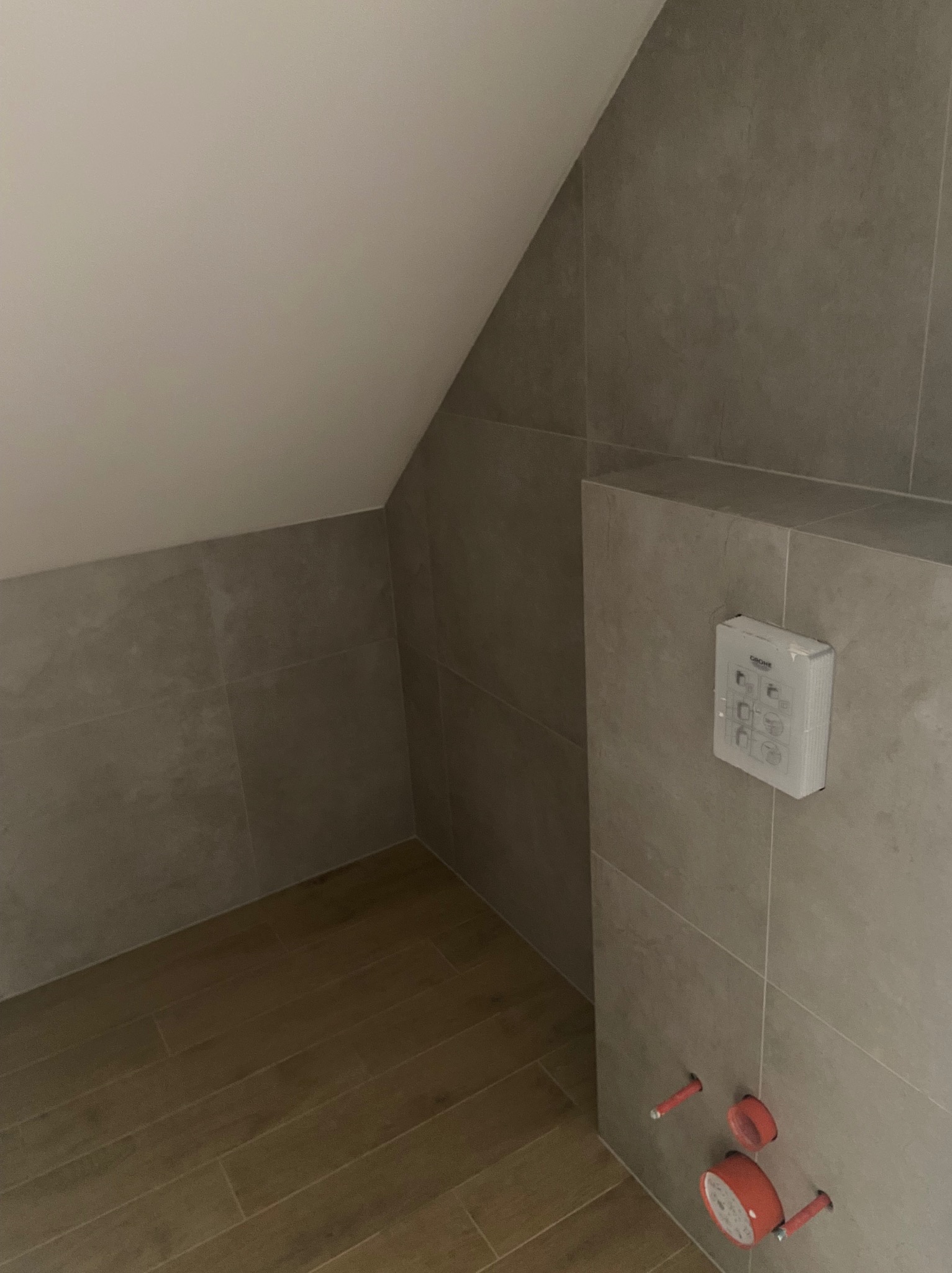 Szara łazienka na poddaszu z drewnopodobną podłogą. Widoczna instalacja sanitarna i przycisk spłukujący.