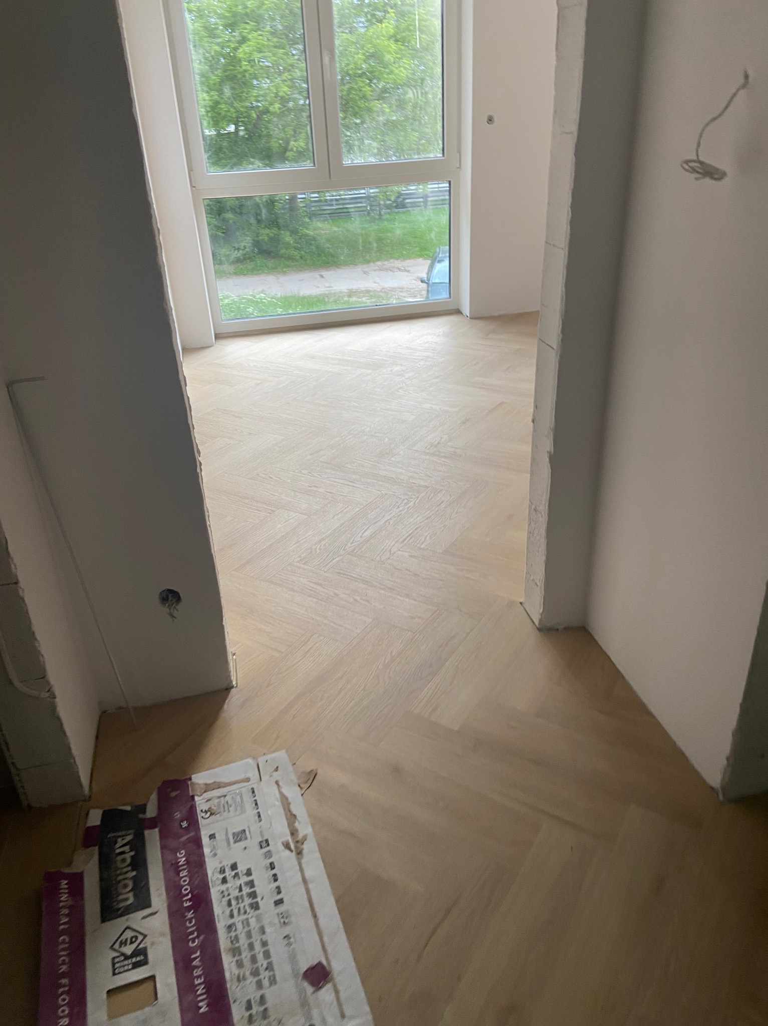 Jasny parkiet ułożony we wzór jodełki francuskiej w nowym wnętrzu. Widoczne kartony po panelach podłogowych. Świeżo wykończone ściany i okno z widokiem na zieleń.