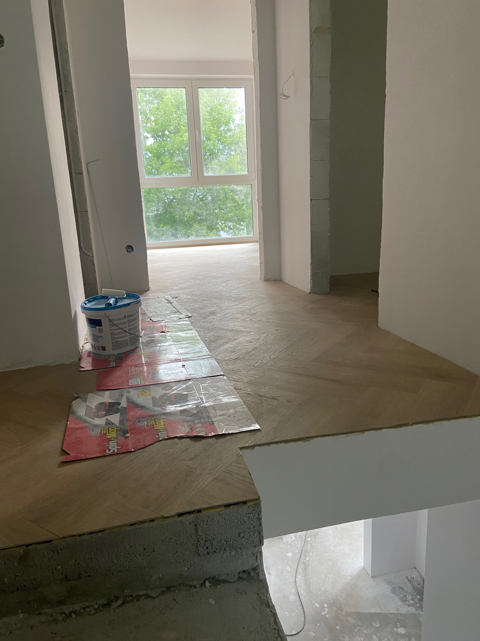 Ukończone układanie parkietu w jodełkę w jasnym wnętrzu, widoczny fragment schodów i wiadro z farbą zabezpieczone kartonami. Przez okno widać zieleń.