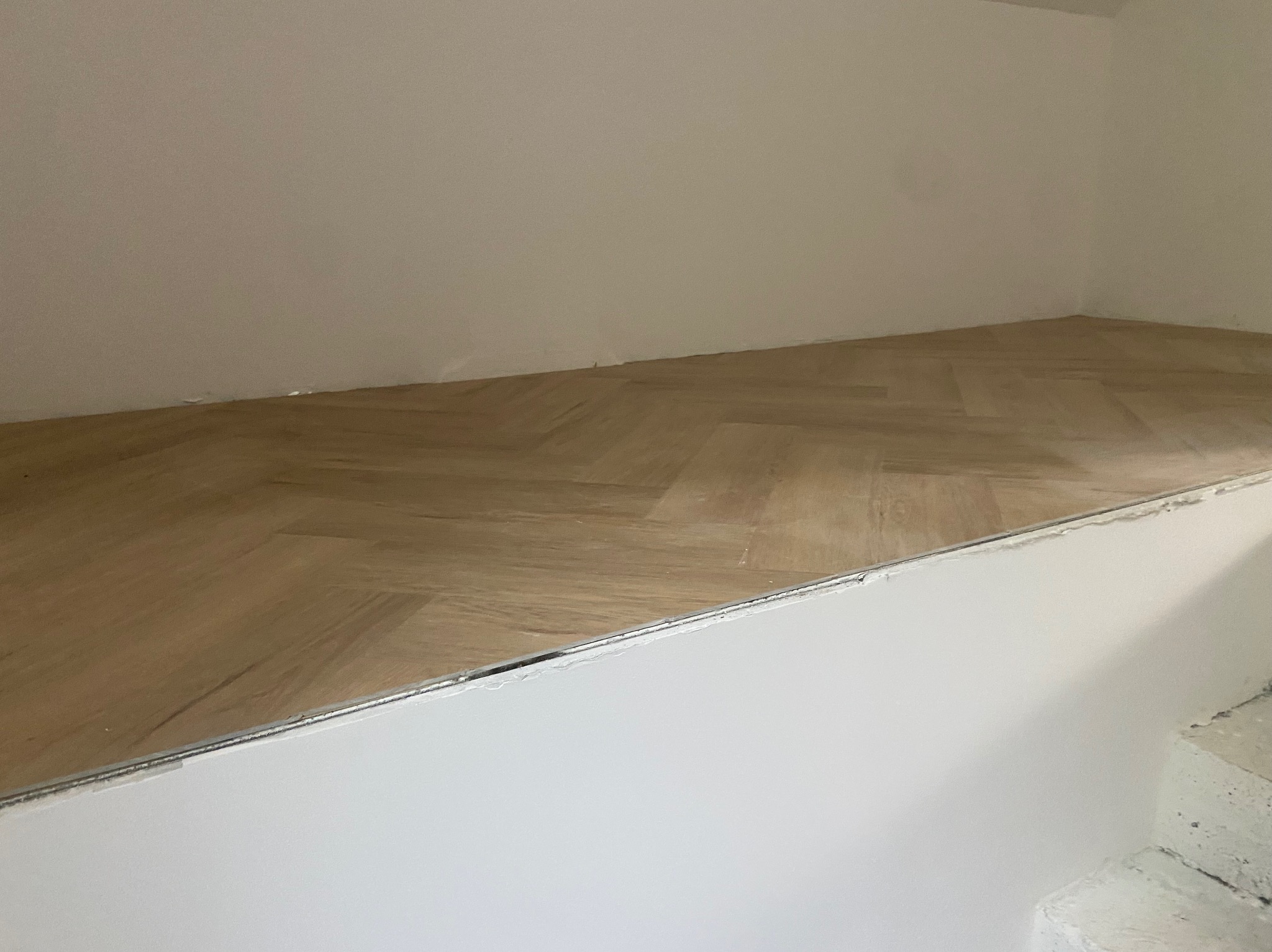 Ukończona podłoga z parkietu ułożonego w jodełkę, widoczna z perspektywy podłogi. Jasne drewno kontrastuje z białymi ścianami i schodami.